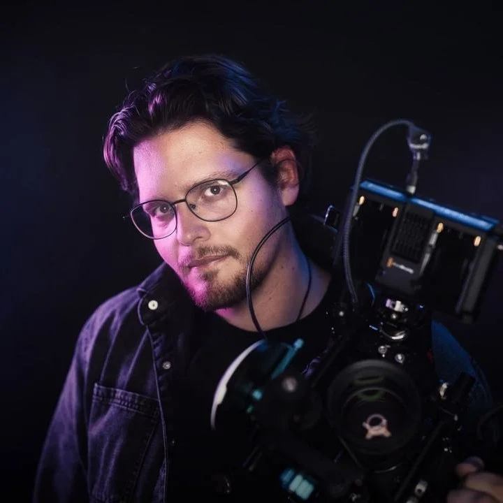 A man with glasses holding a professional camera, posing against a dark background.