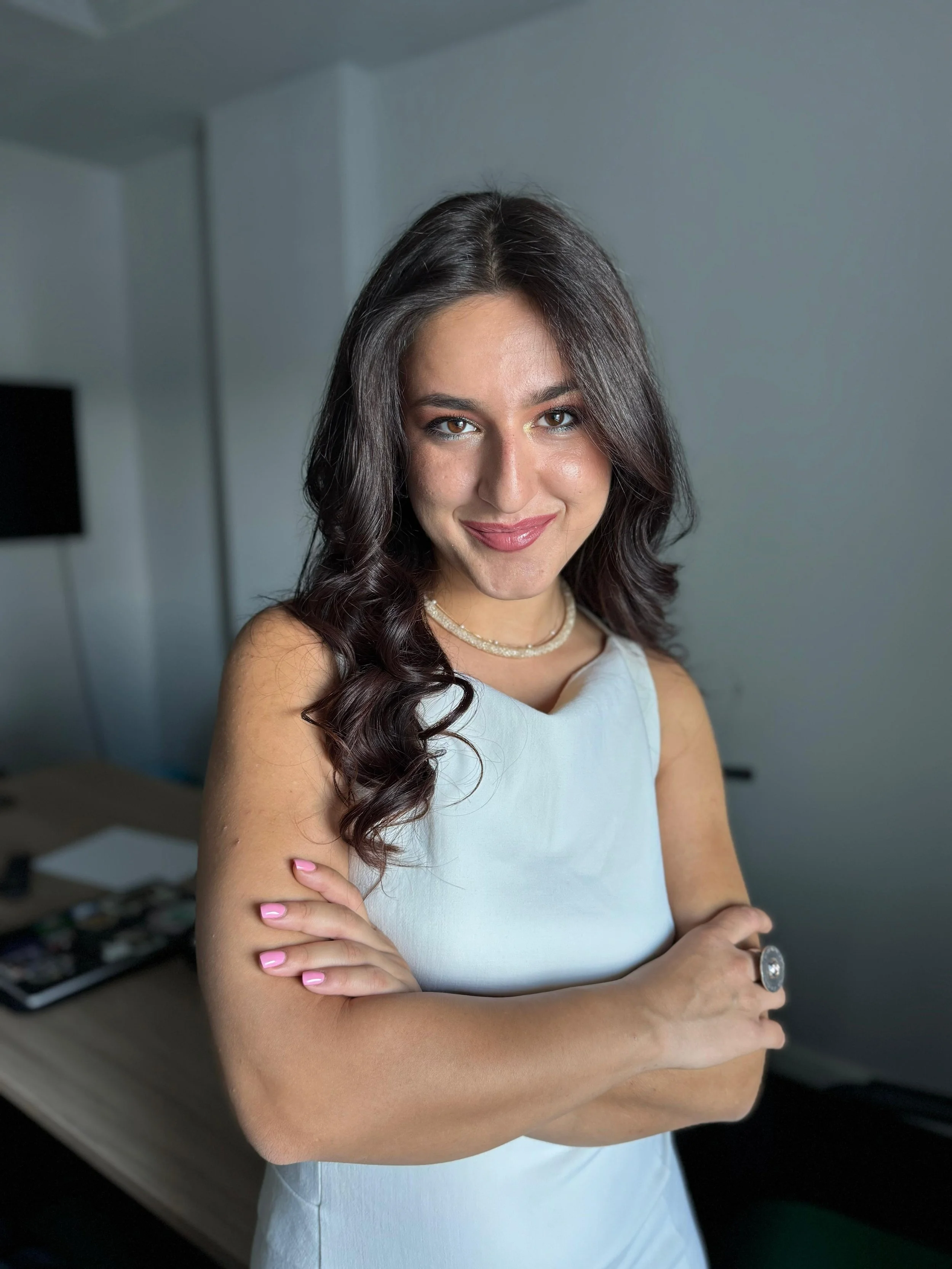 A woman with long, dark, curly hair wearing a white sleeveless top, a pearl necklace, and a ring, standing indoors with arms crossed, smiling at the camera.