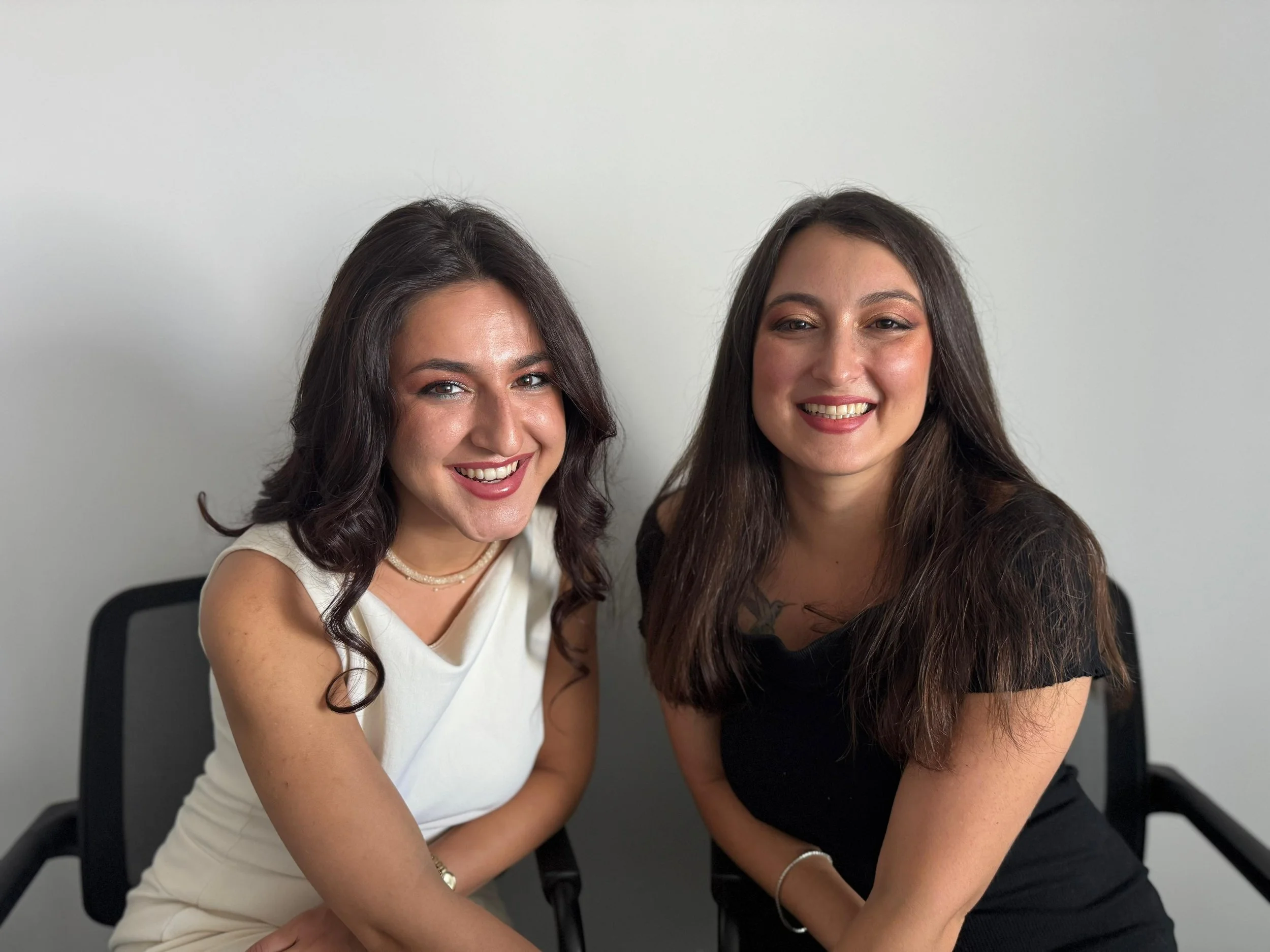 Two women sitting side by side, smiling at the camera, in front of a plain white wall