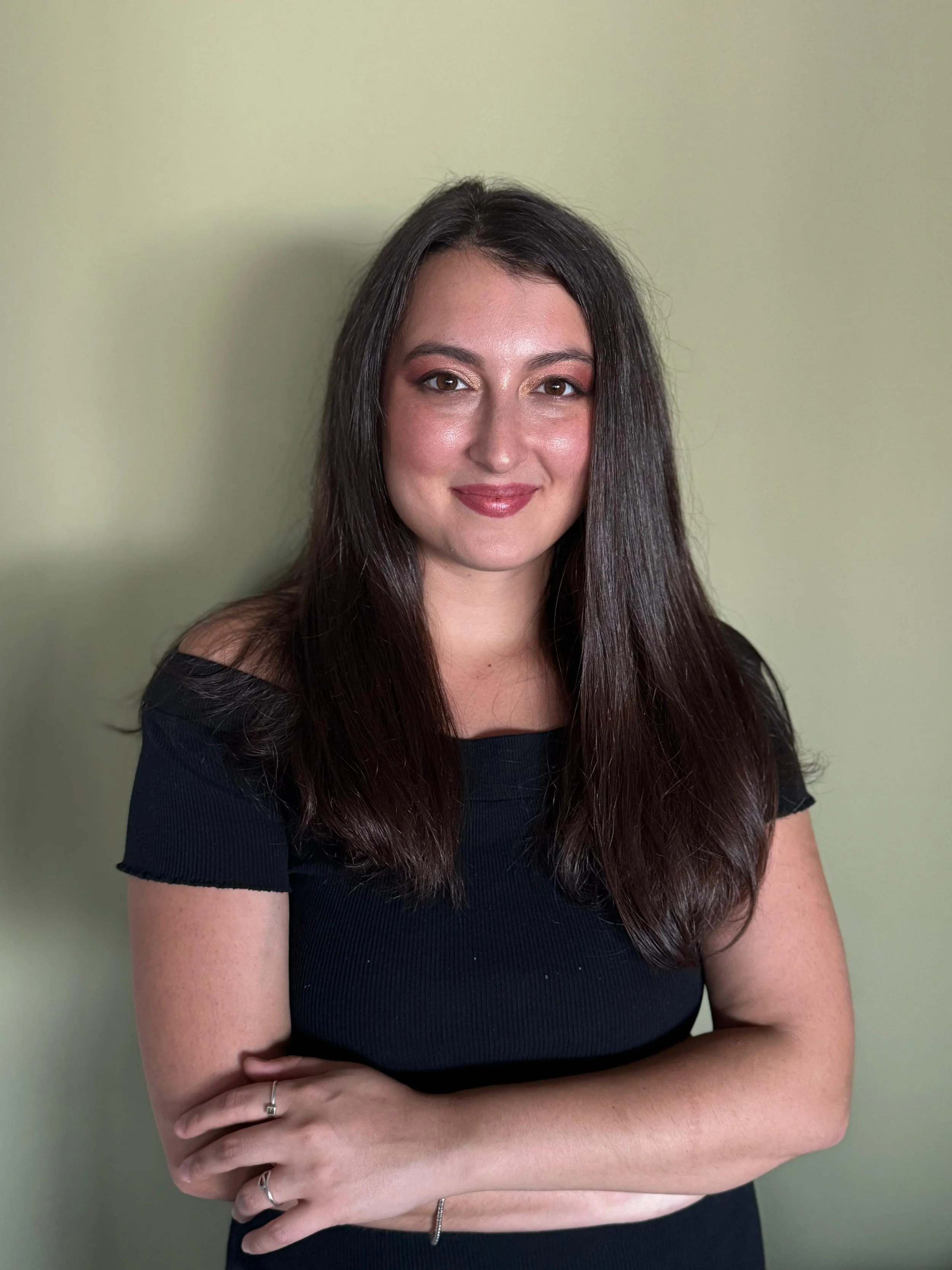 A young woman with long dark hair and a black off-shoulder top, smiling with arms crossed, standing against a light green wall.