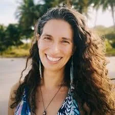 Woman with long curly hair smiling outdoors, wearing a patterned top and earrings, with trees and sunlight in the background.