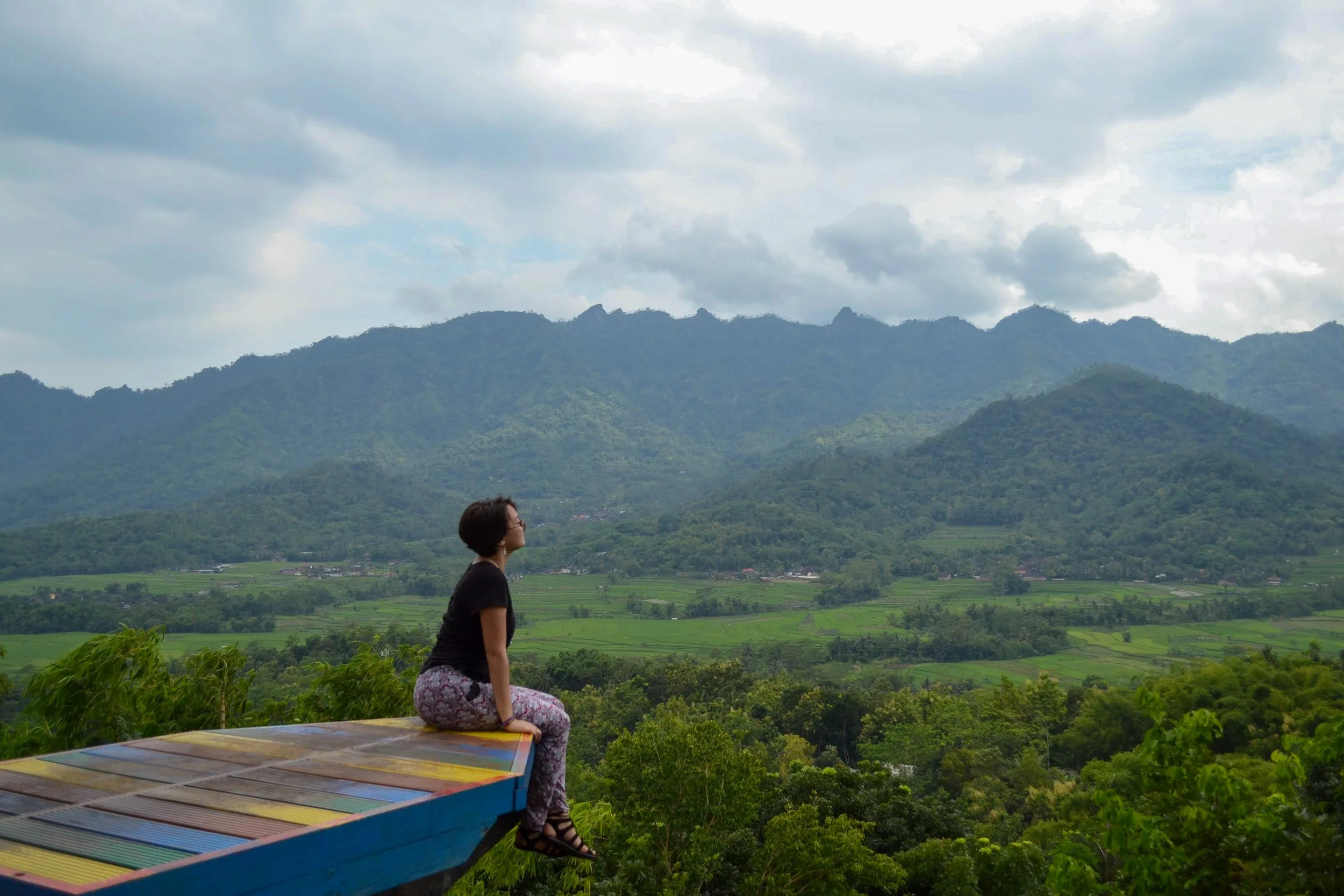 A woman sitting on a colorful wooden ledge overlooking green mountains and a cloudy sky.