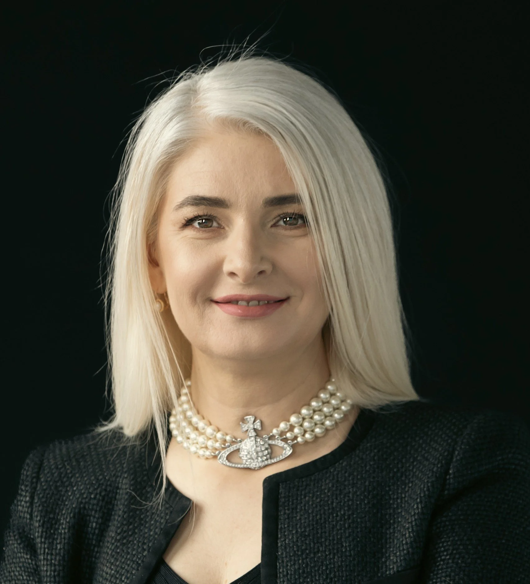 Portrait of a woman with platinum blonde hair wearing a pearl necklace with a jeweled cross pendant, black textured jacket, and subtle makeup, against a dark background.