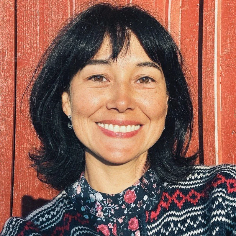 A smiling woman with shoulder-length dark hair, wearing earrings and a patterned sweater in front of a wooden wall.