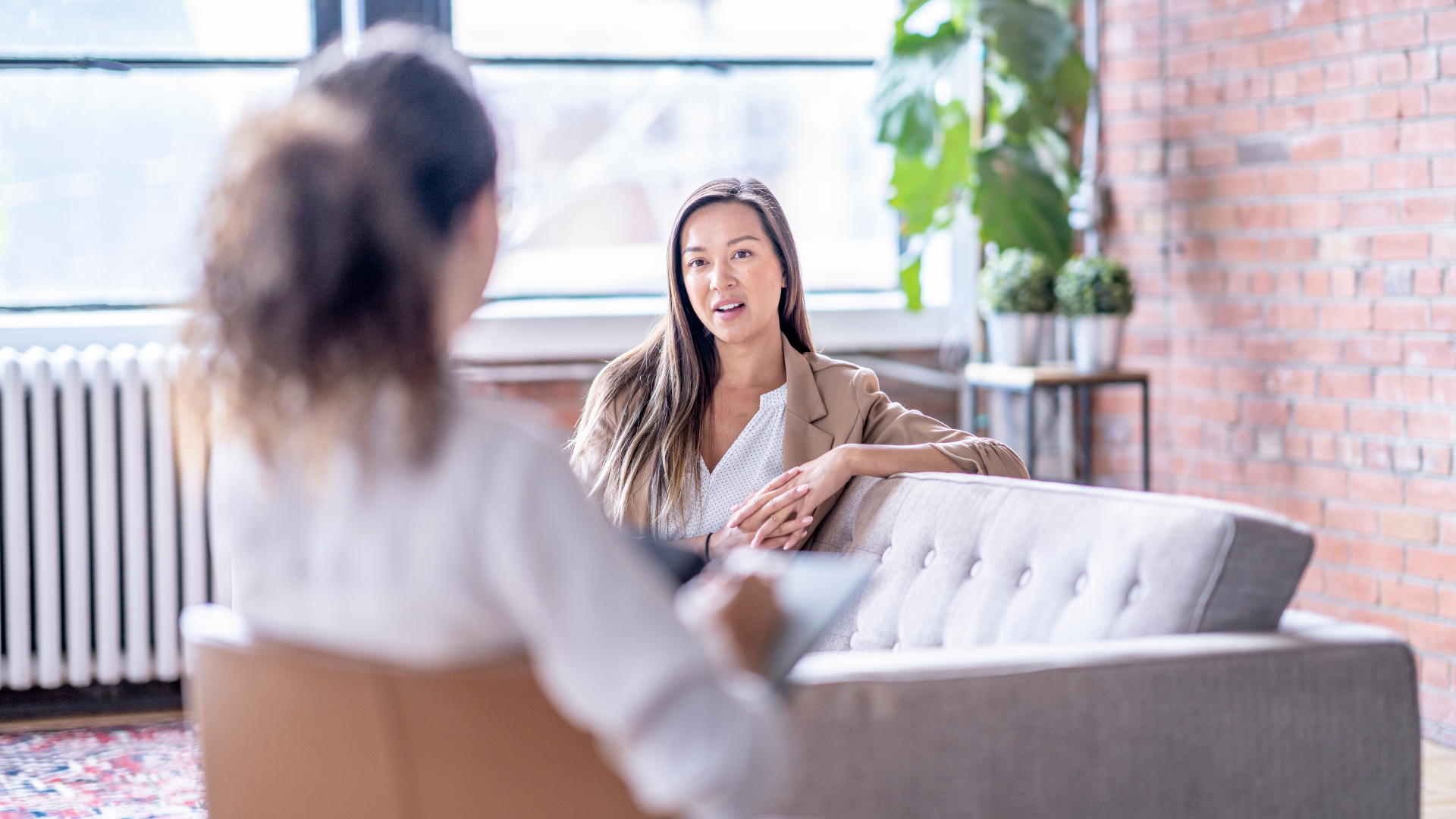 Two women in a conversation in a bright, modern room with large windows, a brick wall, and potted plants.