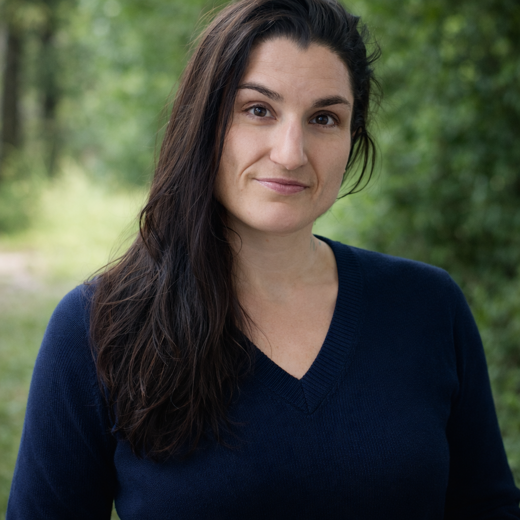 A woman with long, dark hair and a slight smile, wearing a navy blue sweater, standing outdoors with blurred green trees in the background.