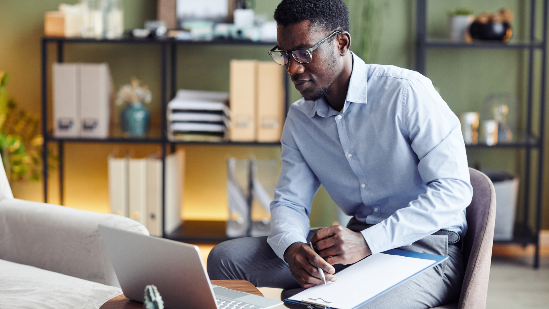 A man wearing glasses in a light blue shirt is sitting at a table, looking down at a device he is holding, with a laptop, notebook, and pen nearby, in a room with shelves and decorative items in the background.