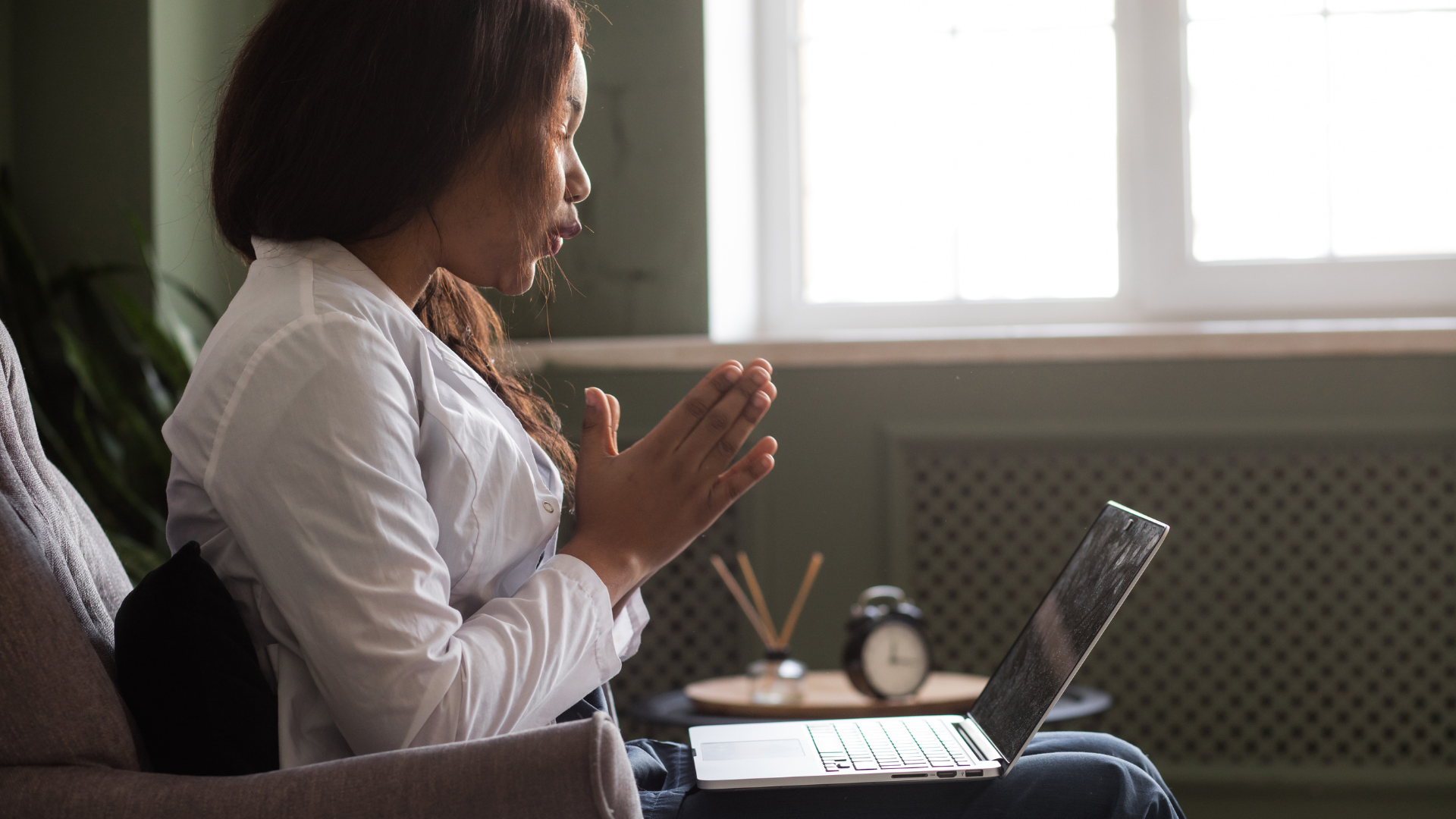 A woman sitting on a couch, wearing a white shirt, praying while looking at her laptop inside a room with a window and a small clock on a table.