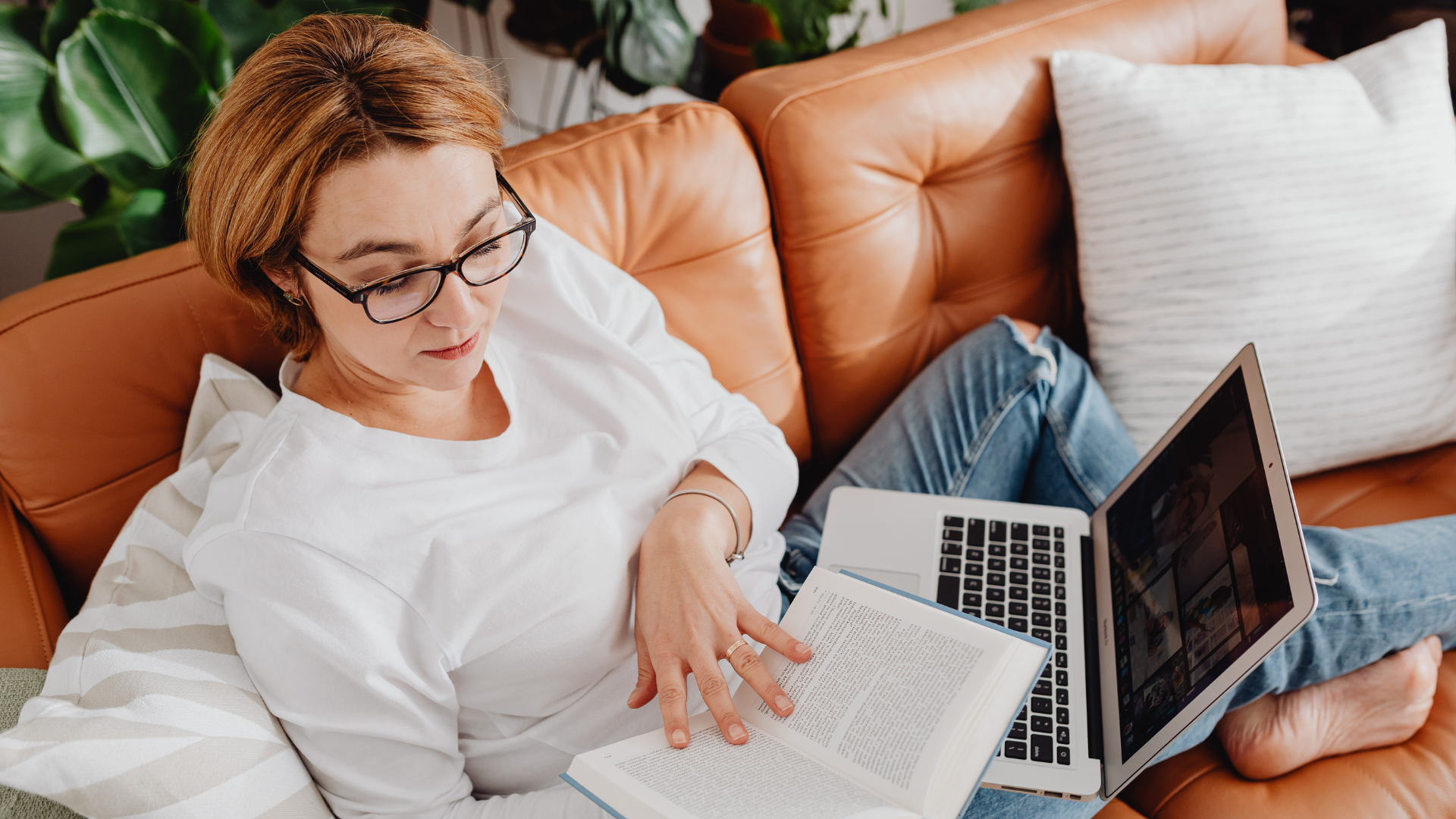 A woman with short brown hair, glasses, wearing a white shirt, is sitting on a brown leather couch. She is holding an open book in one hand and a laptop on her lap, with her legs crossed. Behind her is a large pillow, and in the background, there are green plants.