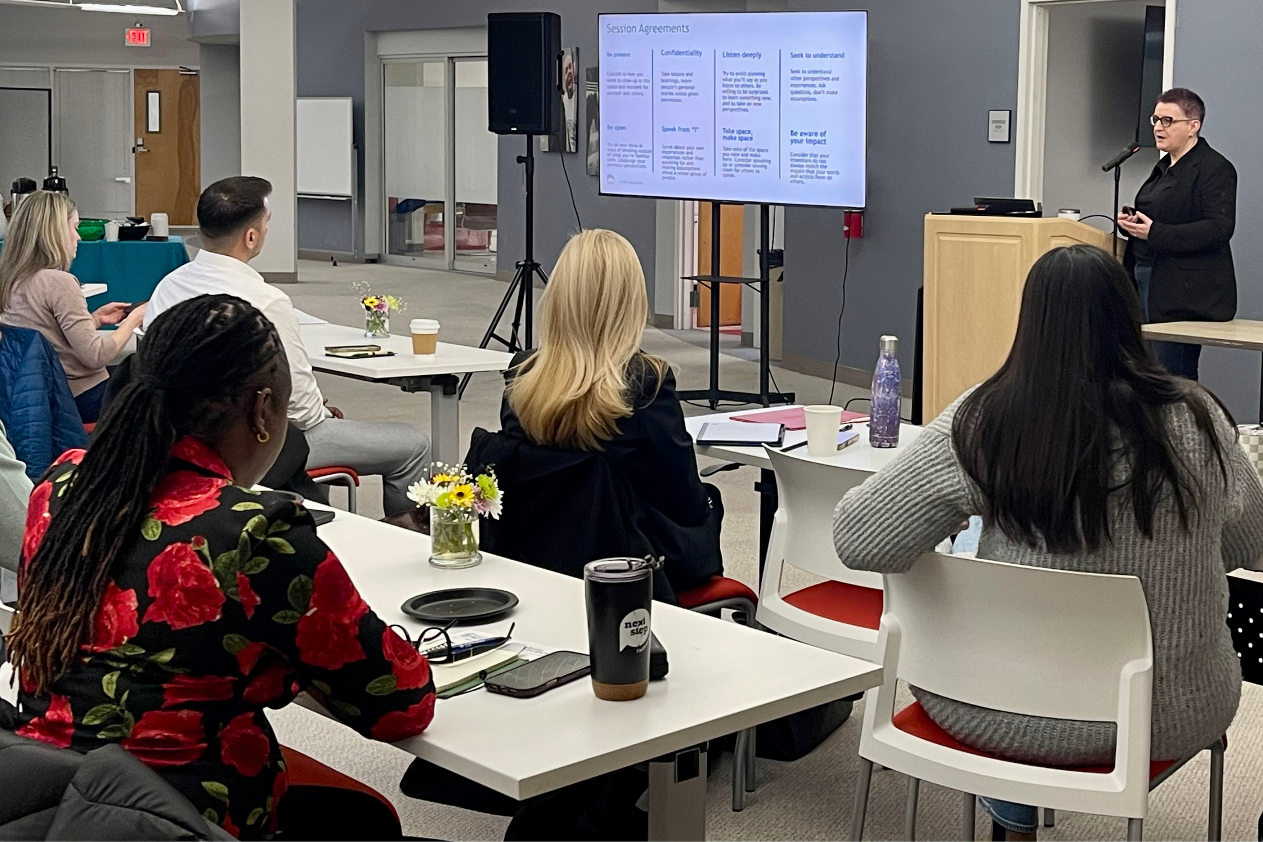 Ciara giving a presentation in a conference room with a large screen displaying session agreements, while attendees sit at tables listening.