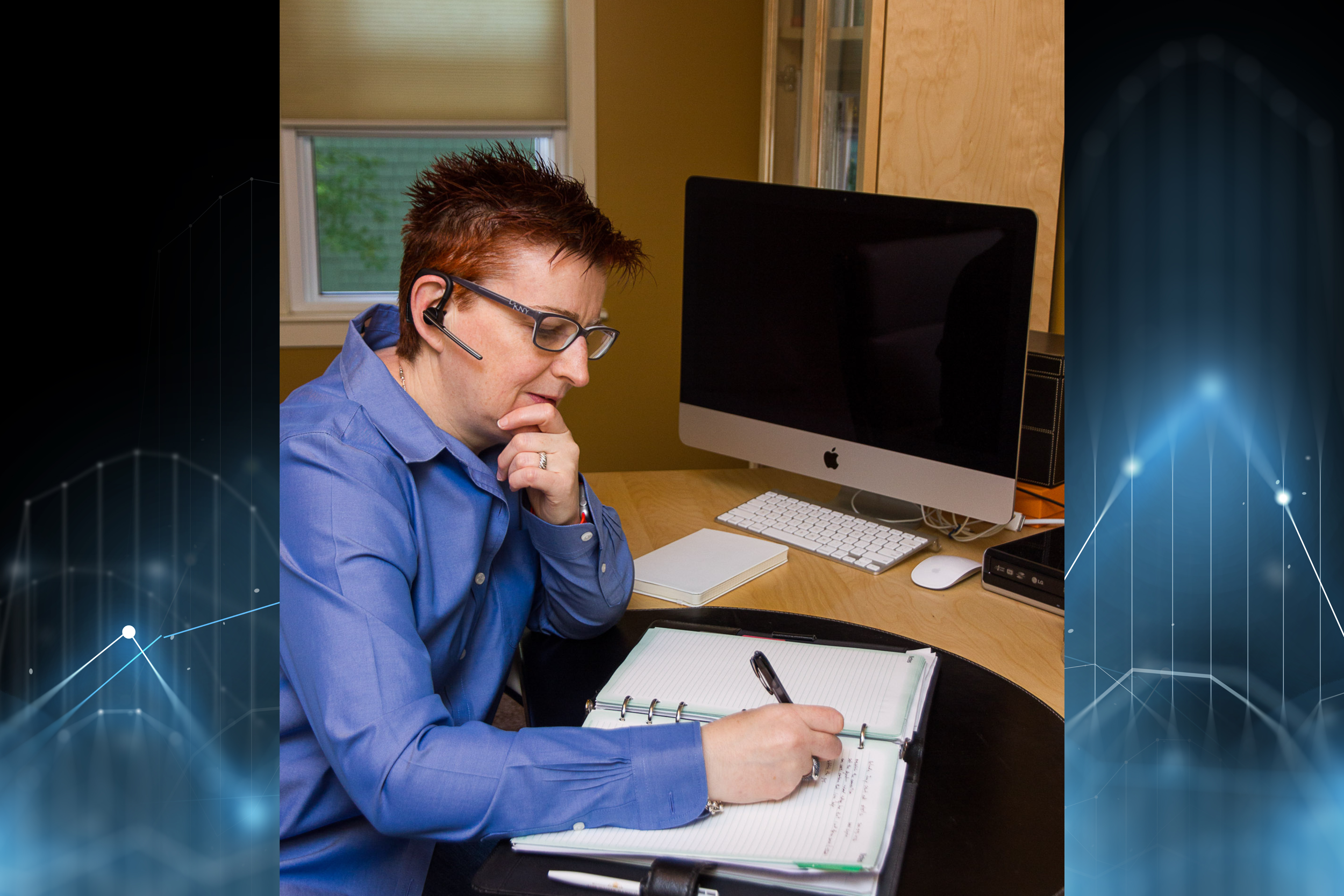 Ciara with short spiky red hair and glasses is sitting at a desk, writing in a notebook with a pen. She is wearing a blue shirt and has a headset around her neck. She is conducting analysis for a client.