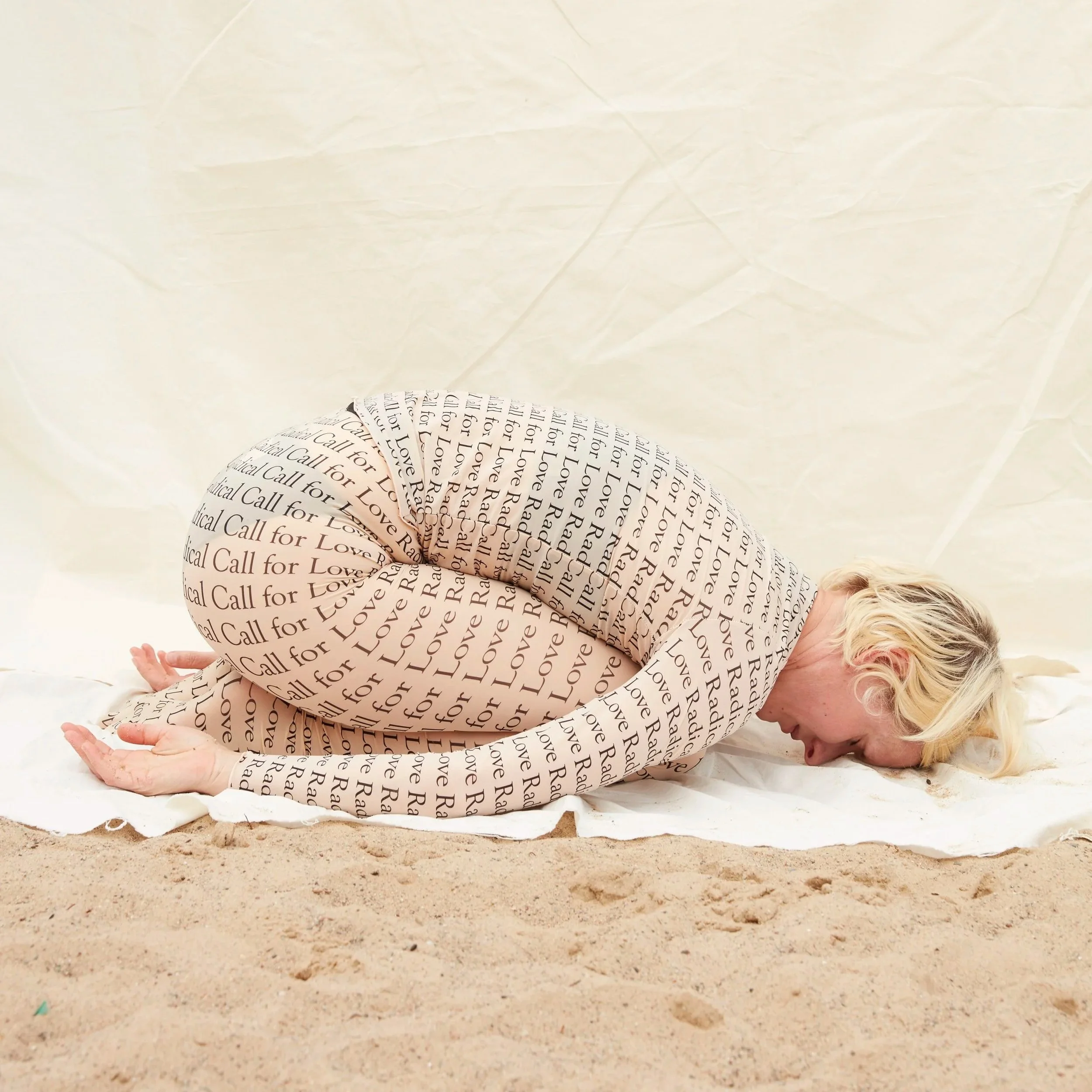 A woman with blonde curly hair wearing a dress with the words 'Love' and 'Call for Love' repeatedly printed on it, lying on her side on a white blanket on the sand with her head bowed and face tilted toward the ground.