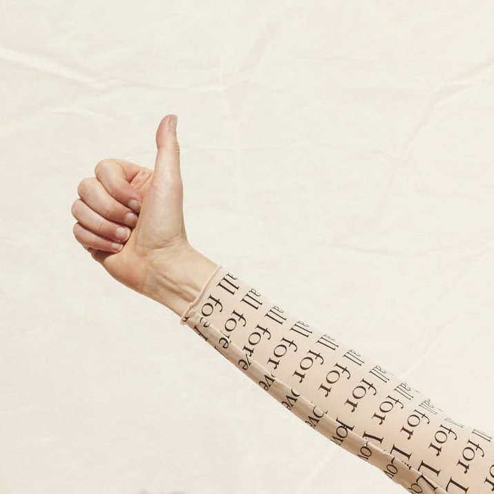 A person's arm with tattooed text, giving a thumbs-up gesture against a plain light background.