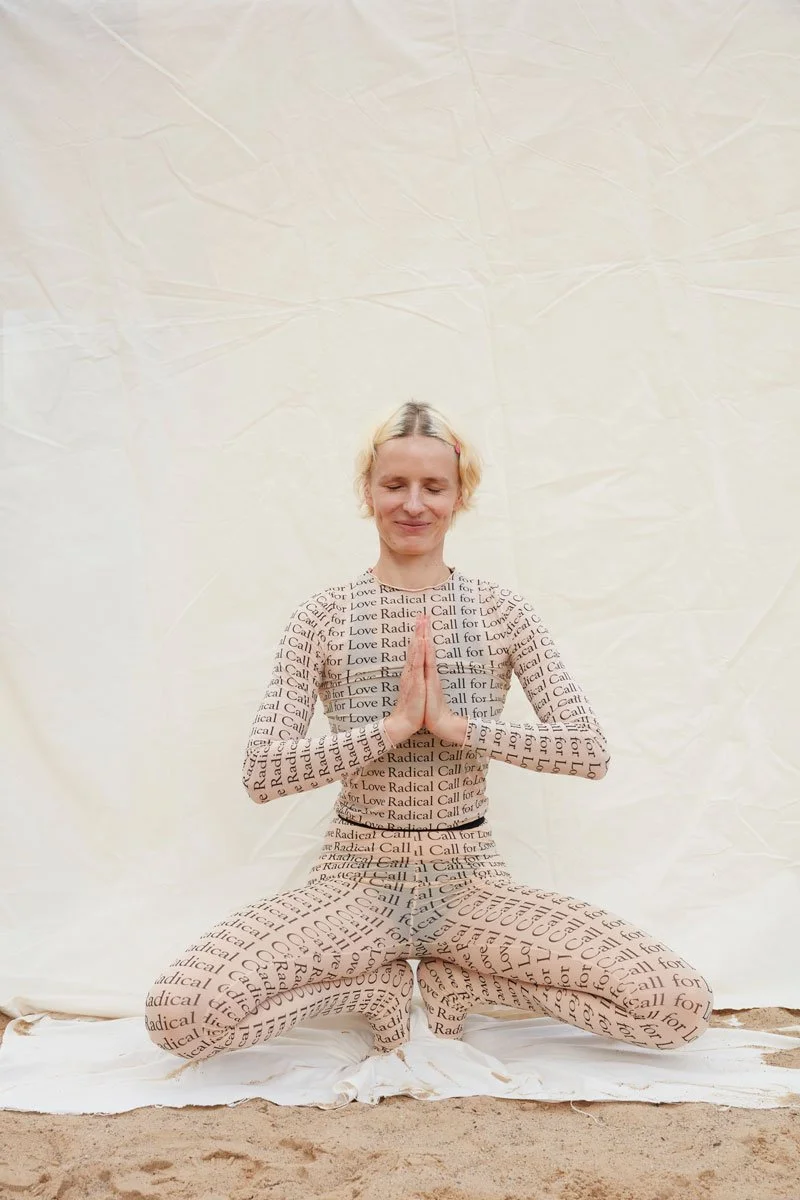 A woman wearing a matching dress and leggings with the words 'Call for Love Radical' is kneeling on the ground with hands pressed together in a prayer position, eyes closed, in front of a plain white backdrop.