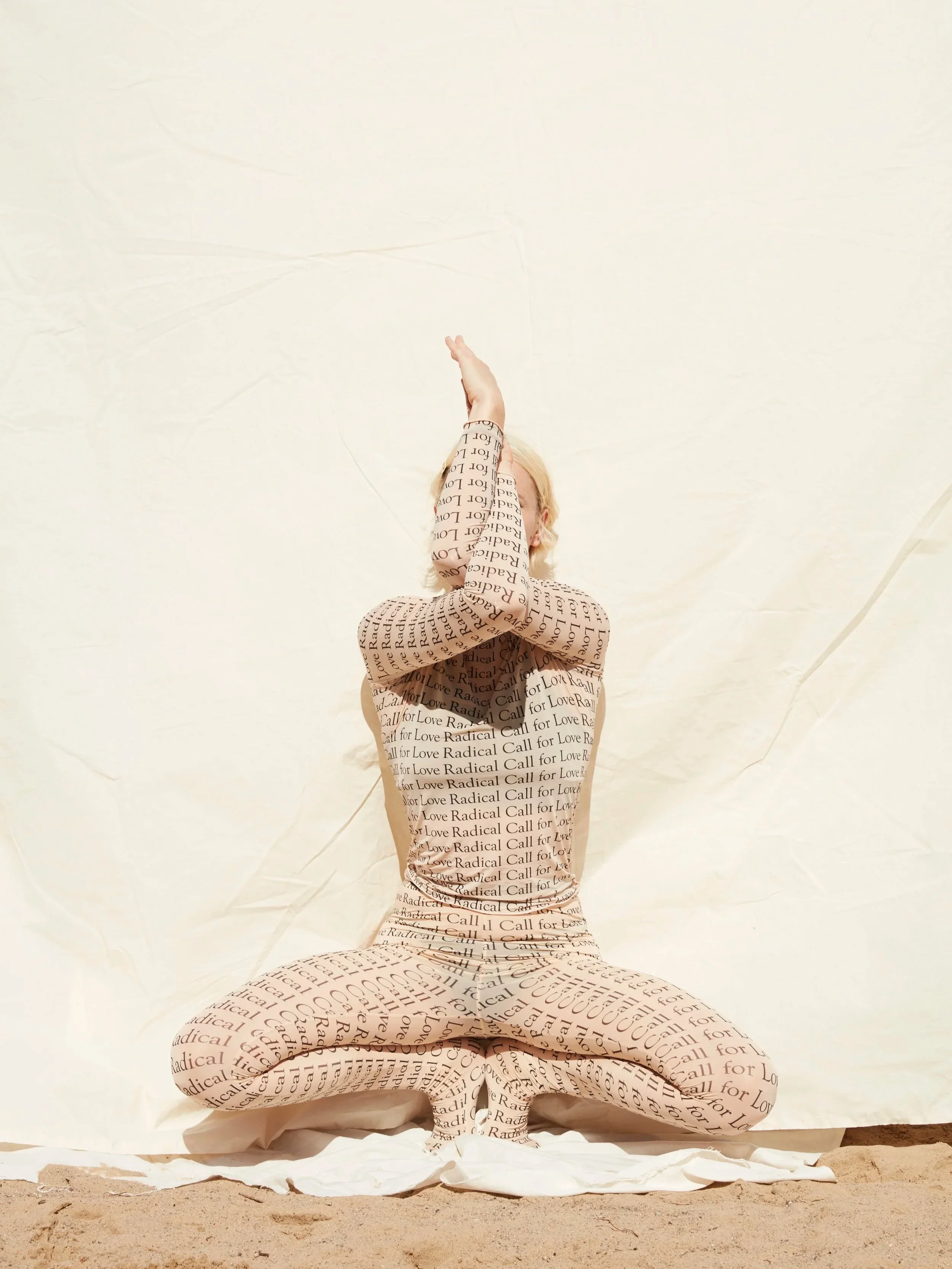 Person in beige and black text-covered outfit kneeling on the ground with arms raised, against a plain light-colored background.