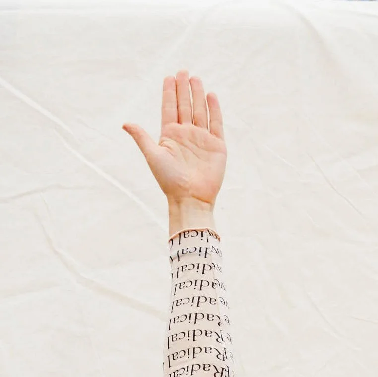 A person's arm raised with the palm facing forward, wearing a white long-sleeve shirt with the word 'Radical' printed repeatedly in black text.