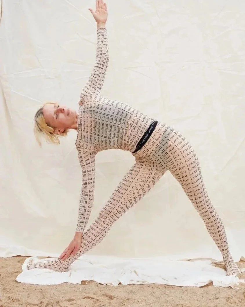 A woman wearing a full-body suit covered in words, performing a yoga pose on a white sheet on the ground against a plain wall.