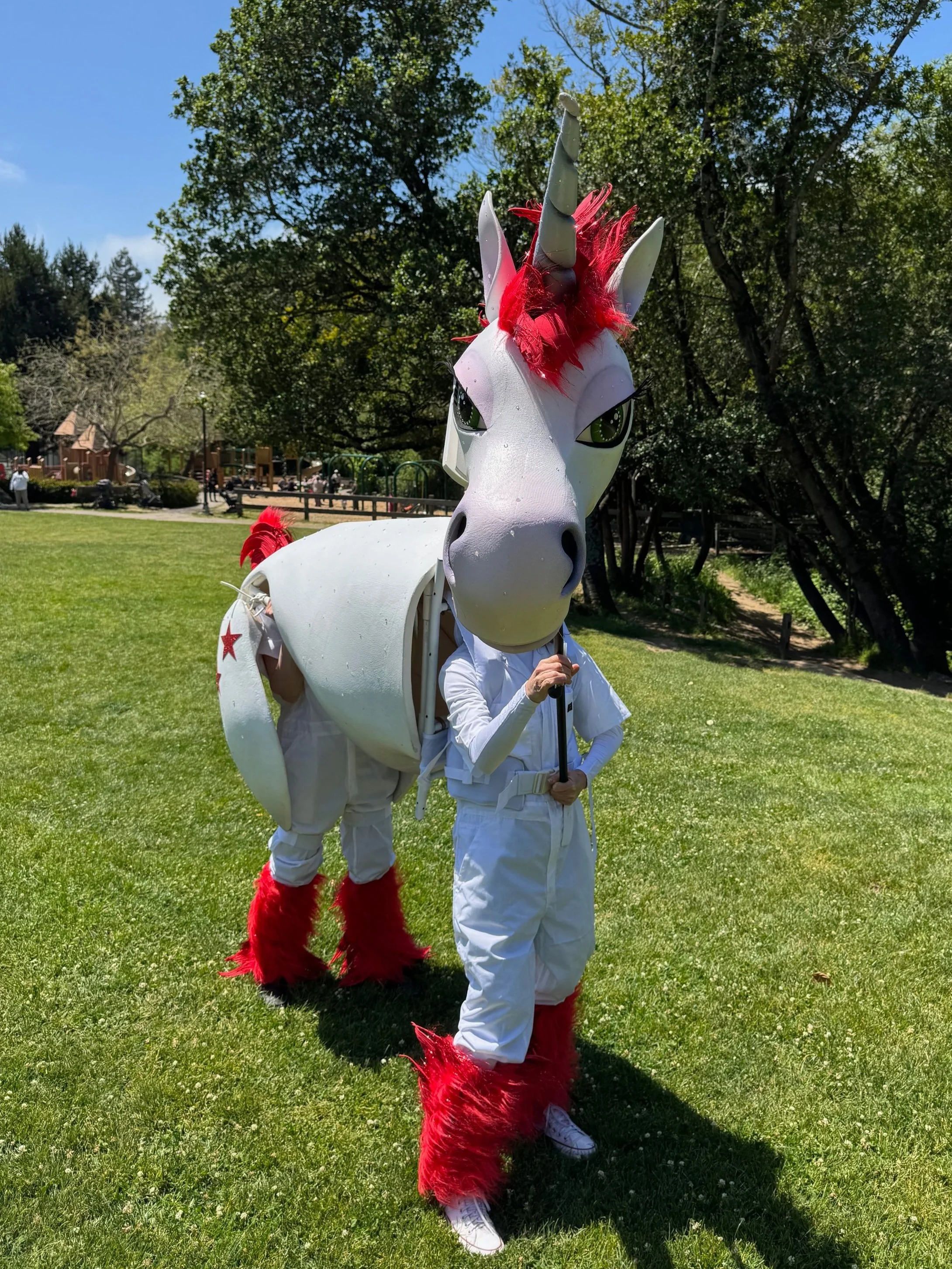 Person dressed in a unicorn costume with a large unicorn head mask, red mane, and furry red leg warmers, standing outdoors on a grassy field during daytime.