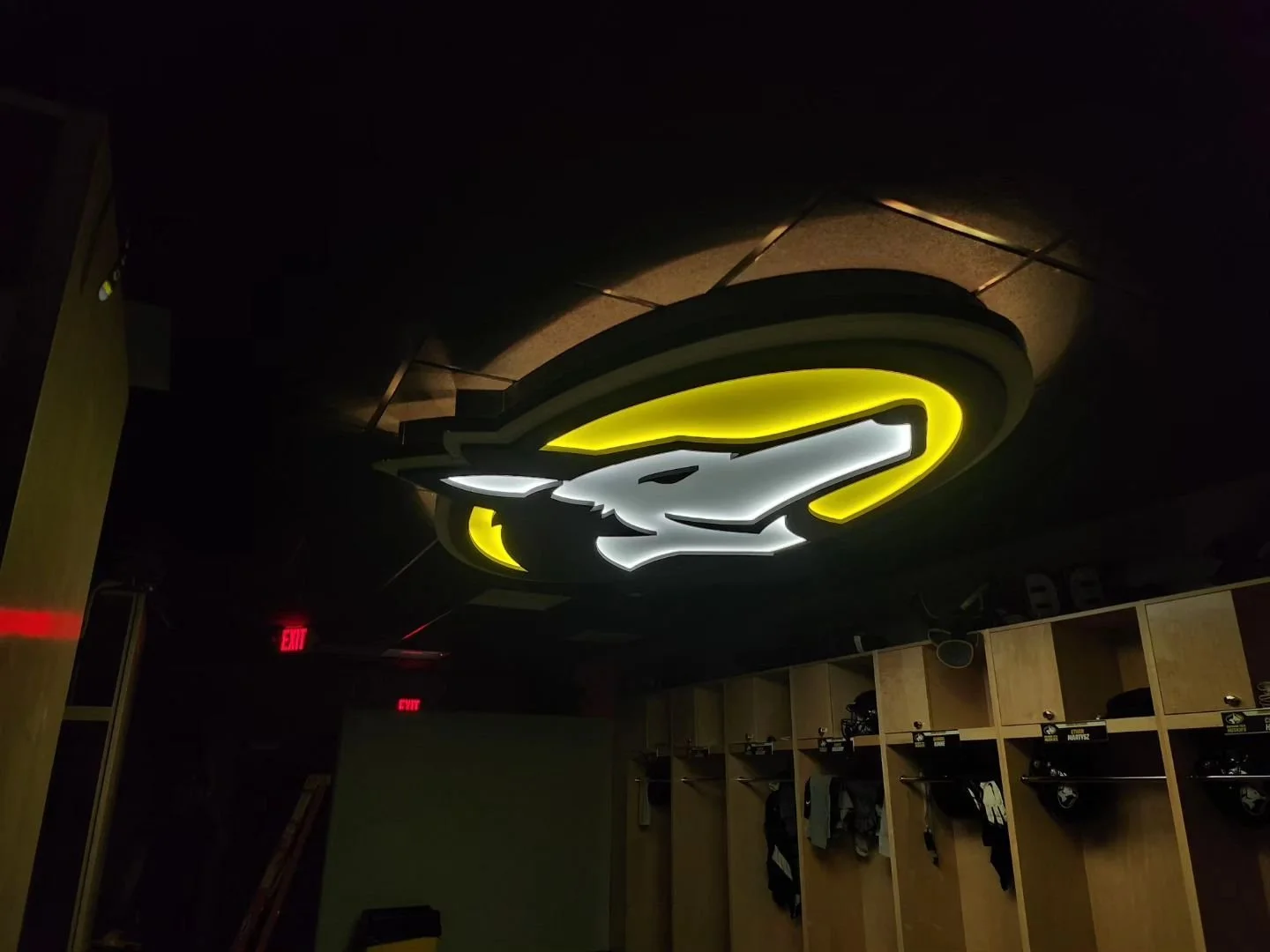 Illuminated logo of the Seattle Seahawks NFL team, featuring a stylized hawk head with yellow, white, and black colors, mounted on a ceiling in a sports locker room with wooden lockers and helmets below.