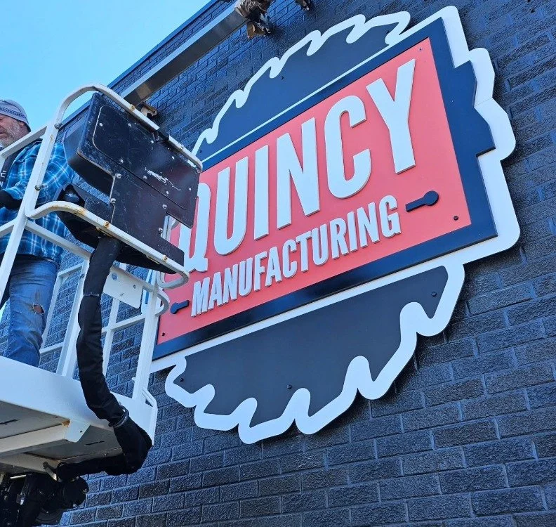 Worker installing a sign that reads "Quincy Manufacturing" on a brick building.