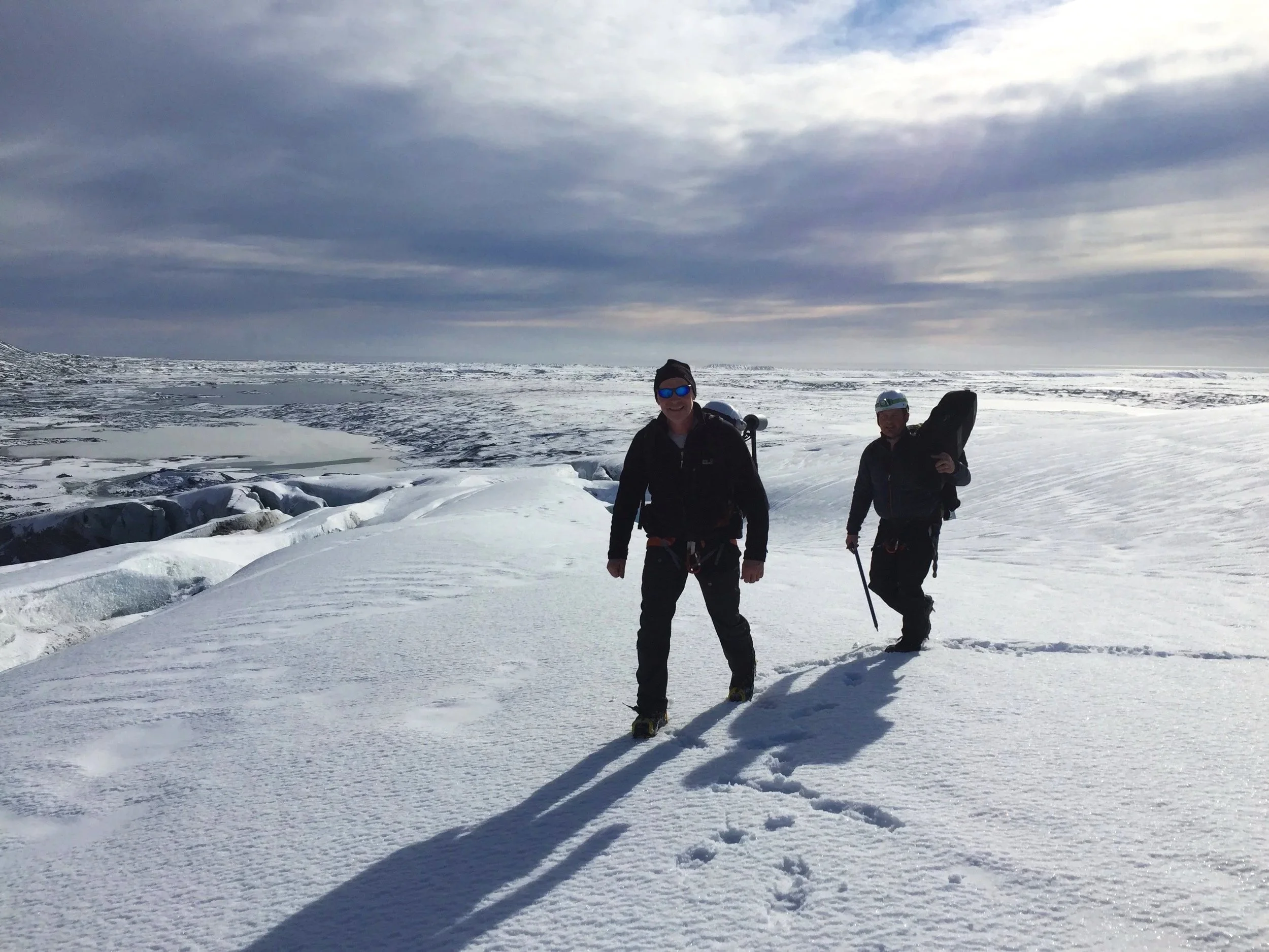 Zwei Menschen in warmer Kleidung und Schneeschuhen wandern auf Schnee in einer arktischen Landschaft mit Eis und Ozean im Hintergrund, bewölktem Himmel.