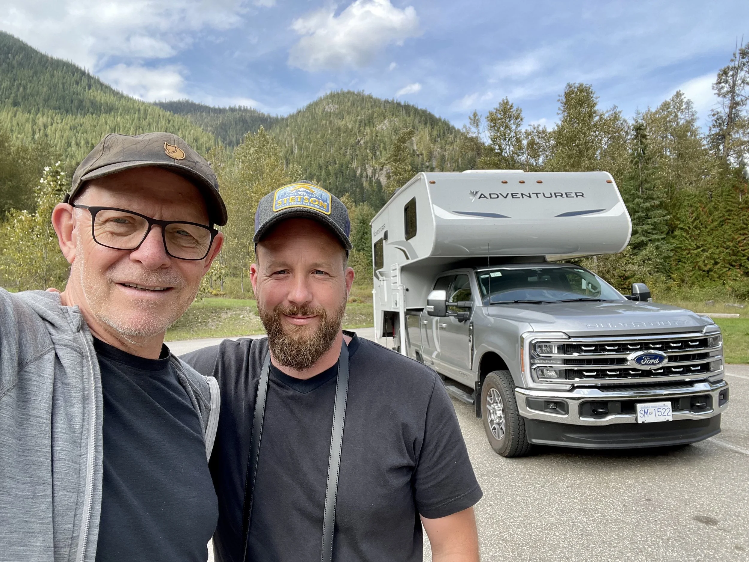 Zwei Männer posieren für ein Selfie vor einem Wohnmobil, im Hintergrund Berge und Bäume, sonniger Himmel.