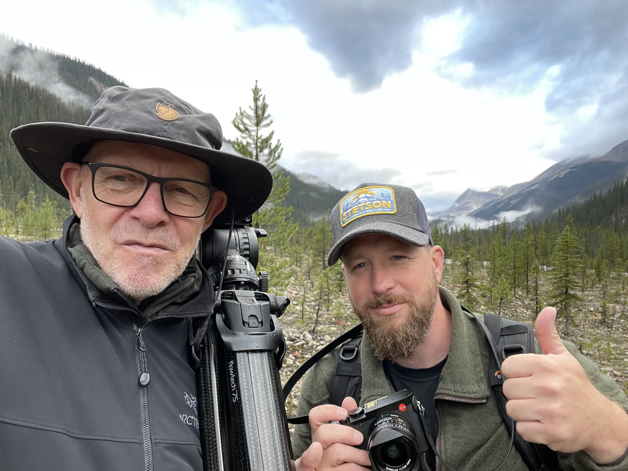 Zwei Männer in Outdoor-Kleidung in einer Berglandschaft mit Bäumen und Wolken, einer mit einer Kamera und der andere gibt Daumen hoch.