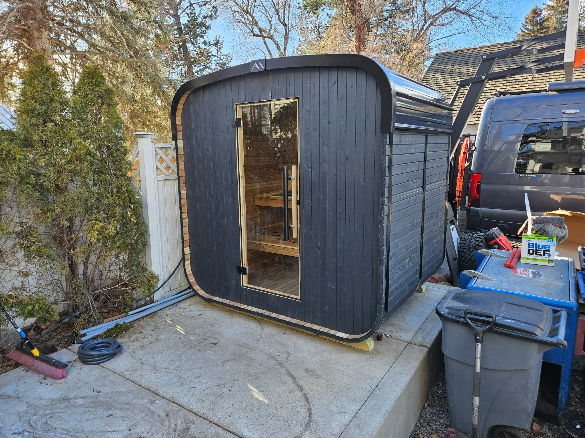 A small black wooden sauna with a glass door is placed on a concrete patio, next to a gray truck, in a backyard with trees and a wooden fence.
