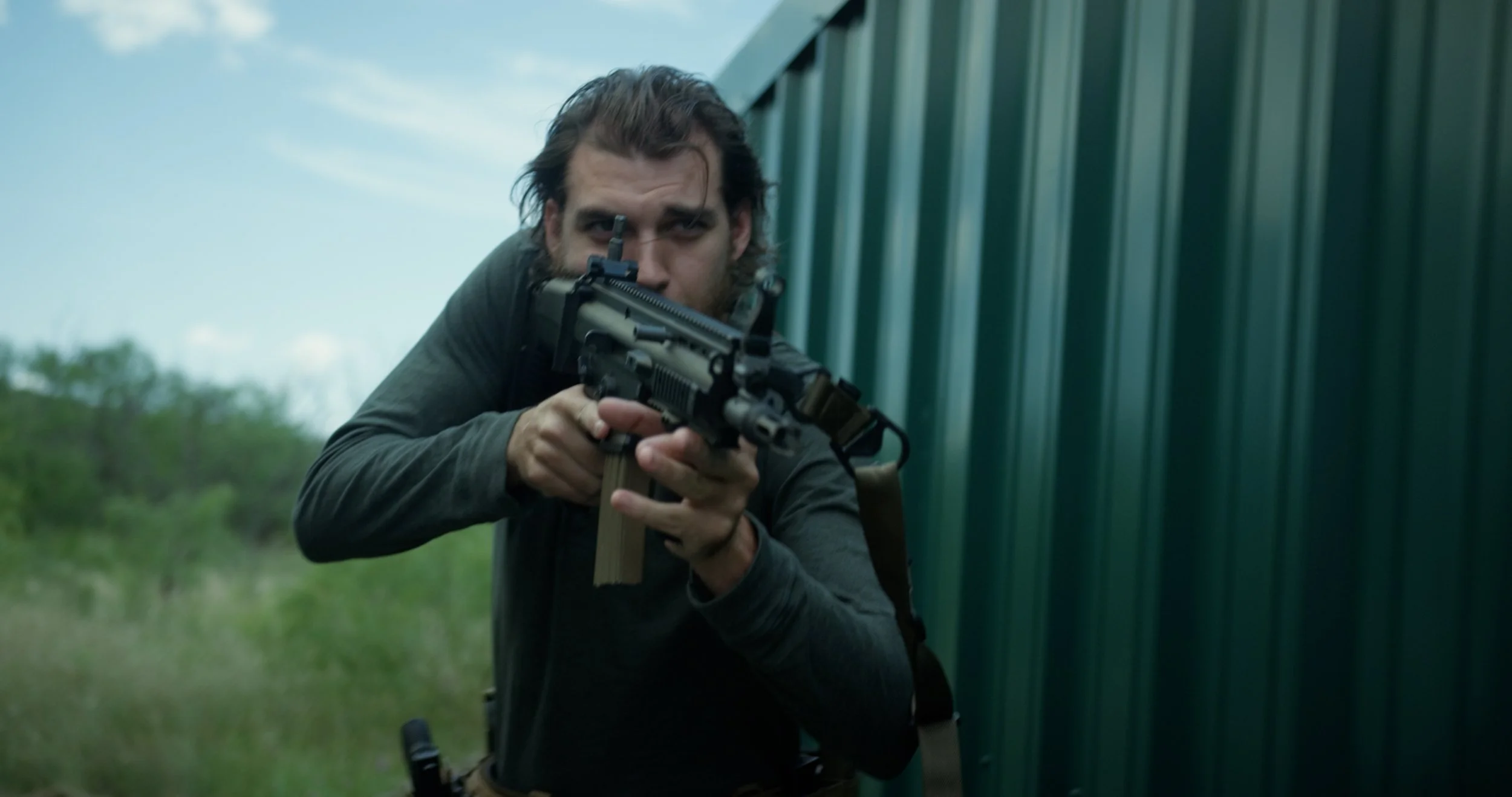 A man with brown hair and beard aiming a handgun with both hands, behind a green metal building, outdoors.