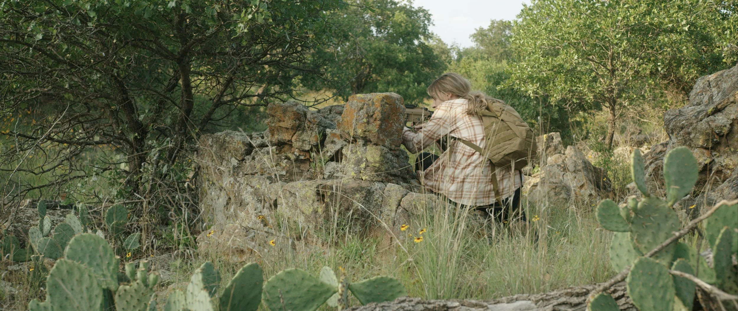 Person with a backpack sitting behind rocks in a desert landscape, surrounded by cacti and shrubs, taking notes.