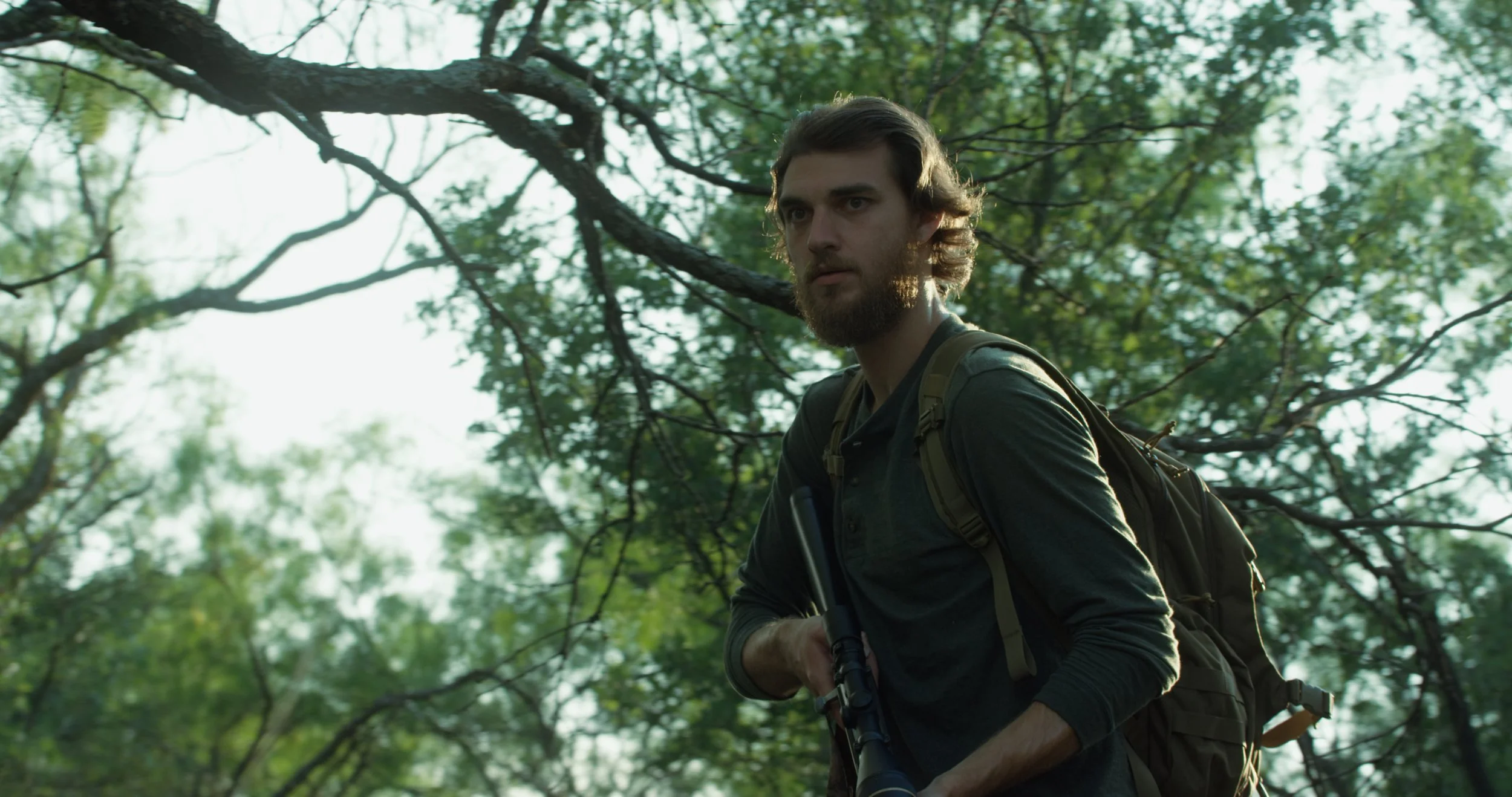 A man with a beard and long hair, wearing a dark shirt, carrying a backpack and holding a rifle, standing in a dense green forest.