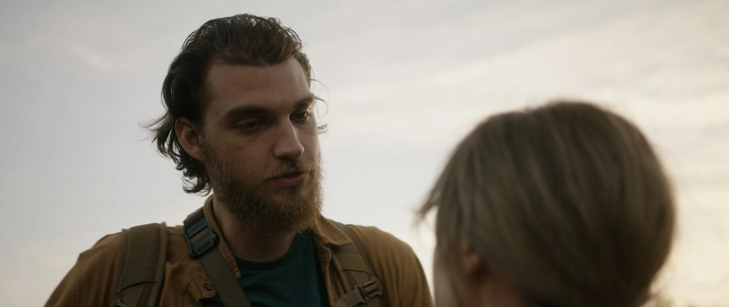 A man with dark hair and a beard looks down at a person with light brown hair, against a cloudy sky background.