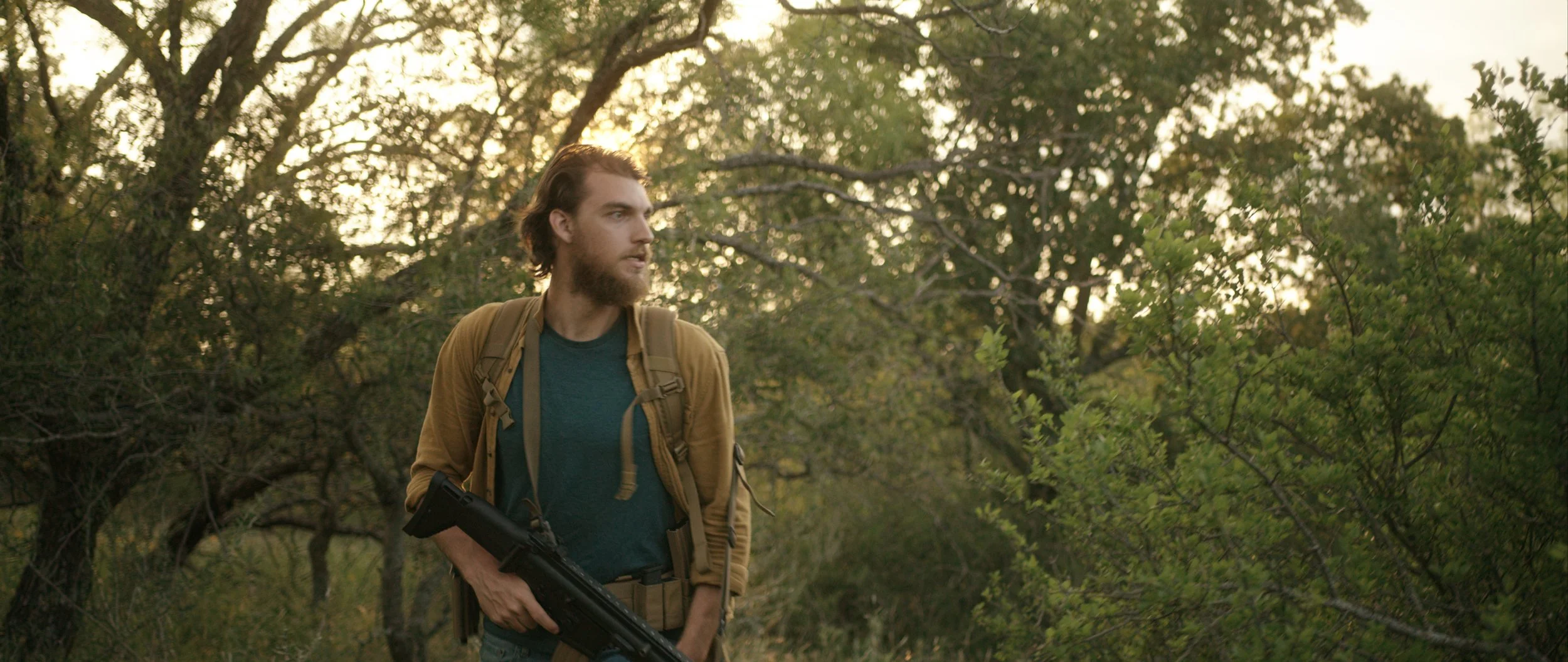 A man with a beard and long hair carrying a gun and a backpack, standing in a wooded area at sunset.