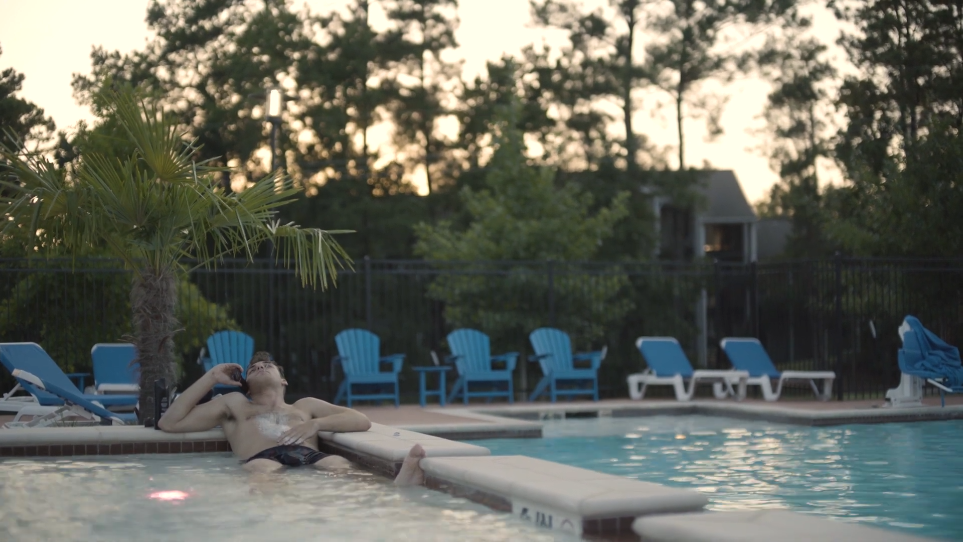 A person is relaxing on the edge of a swimming pool, talking on a cellphone, with lounge chairs and a fenced backyard with trees in the background during sunset.