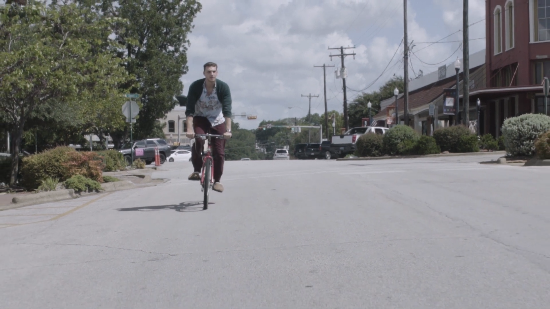 Young man riding a bicycle on an empty street in a small town, with trees, parked cars, and shops in the background.