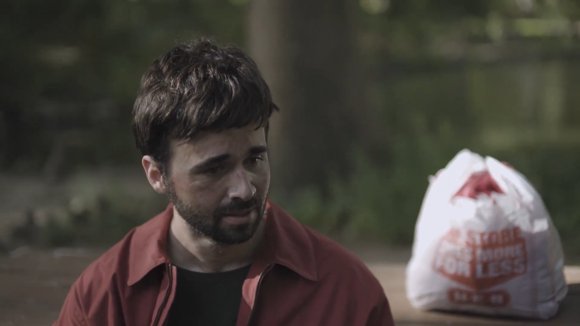 A man with dark hair and beard, wearing a red jacket, sitting outdoors near a water body, with a white plastic bag in the background.