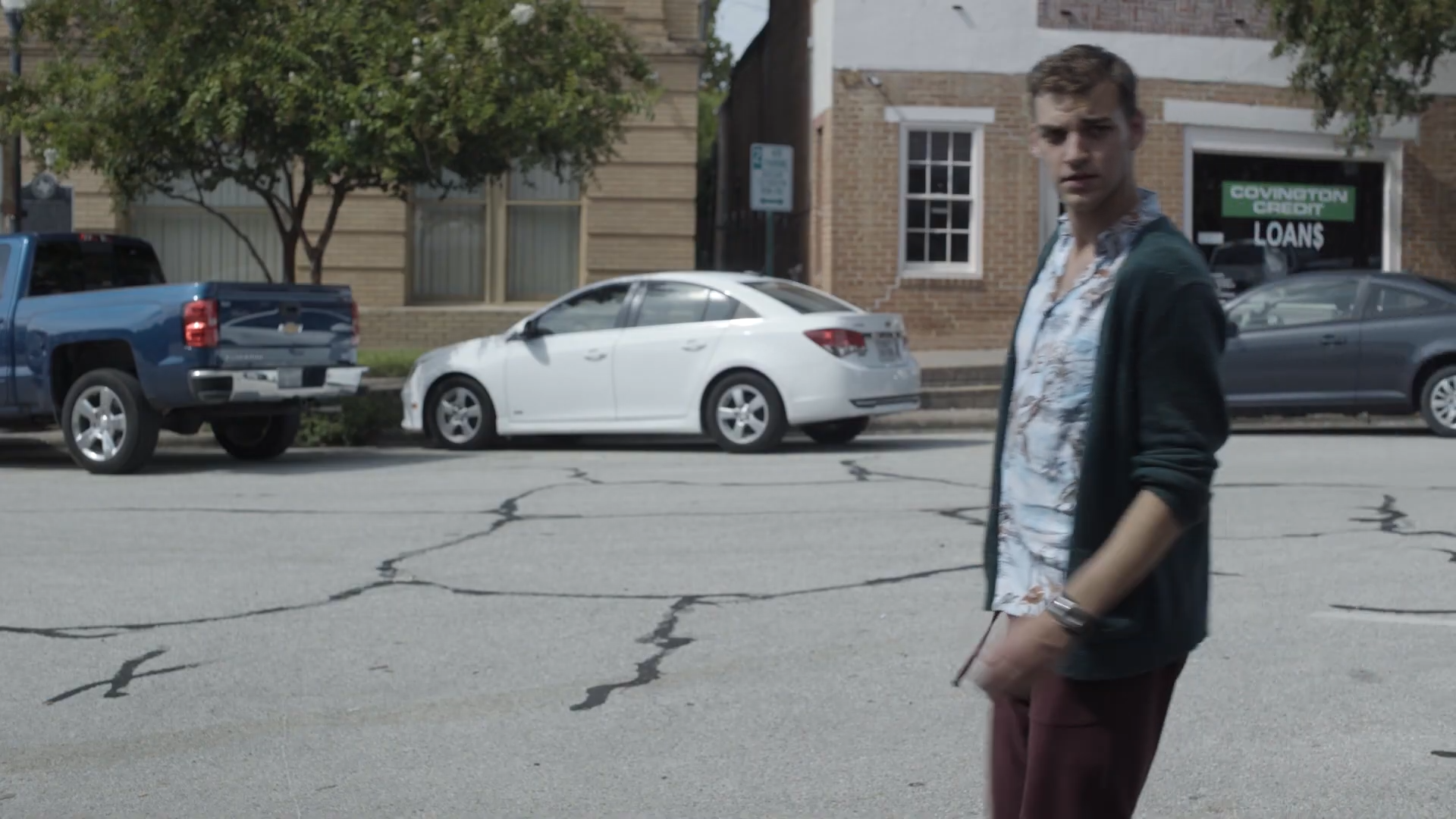 A young man with light brown hair standing on a cracked street, wearing a floral shirt and dark jacket, with parked cars and a brick building in the background.