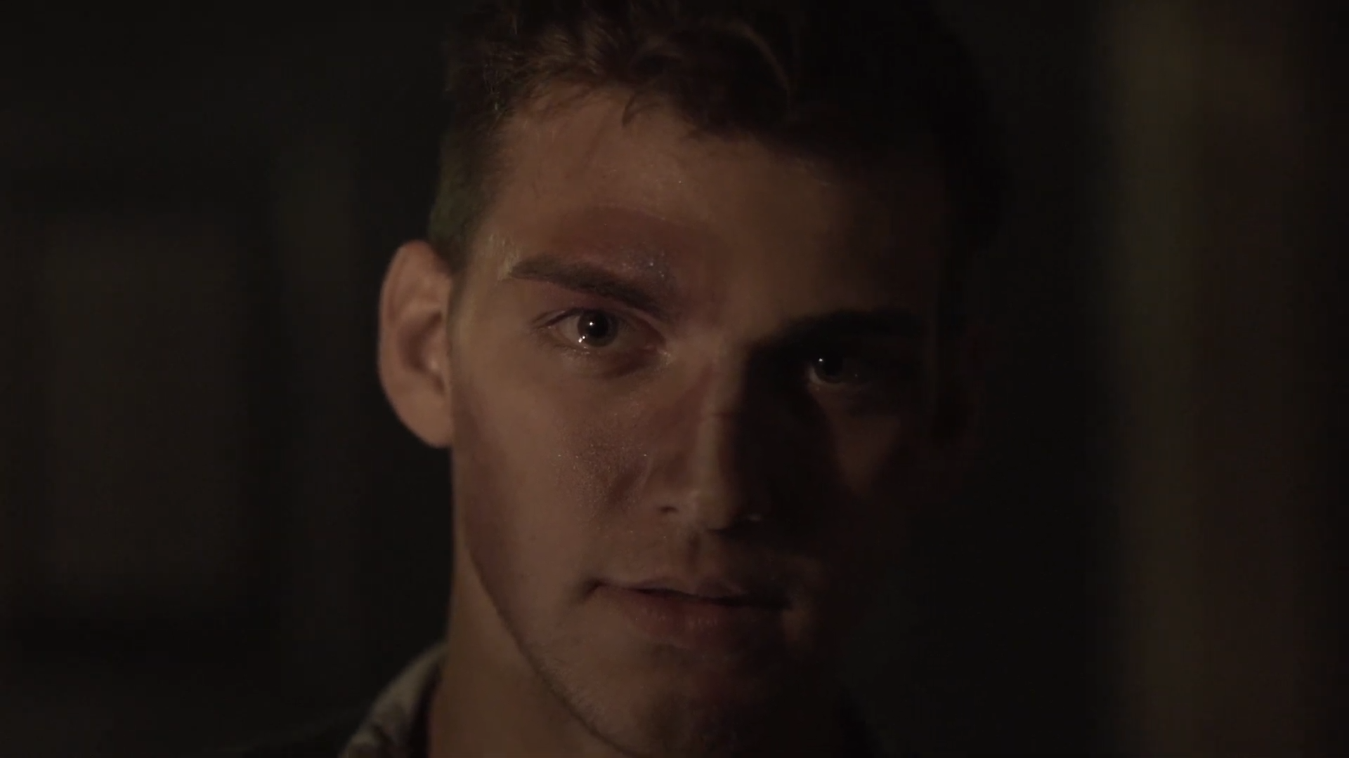 Close-up of a young man's face with a serious expression, dim lighting, and shadows.