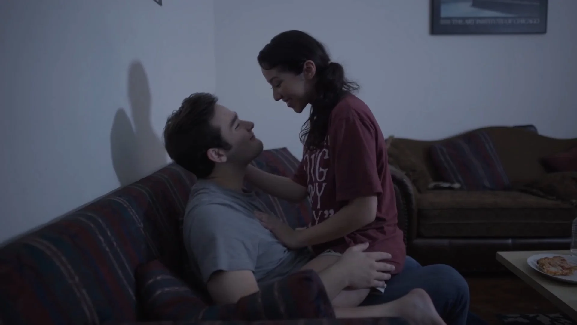 A young man with short dark hair sitting on a couch, smiling and looking up at a young woman with long dark hair, who is leaning over him and smiling, with her hands on his shoulders in a living room setting.