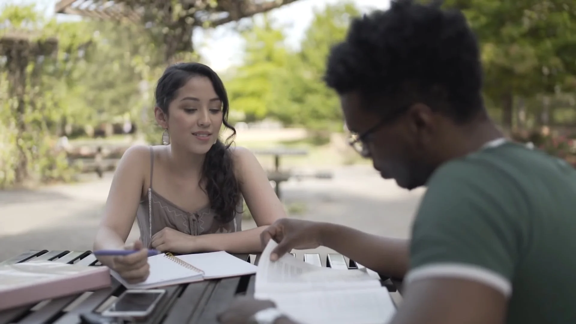 Two young adults sitting at a park bench, studying with books and notebooks in front of them, surrounded by green trees and sunlight.