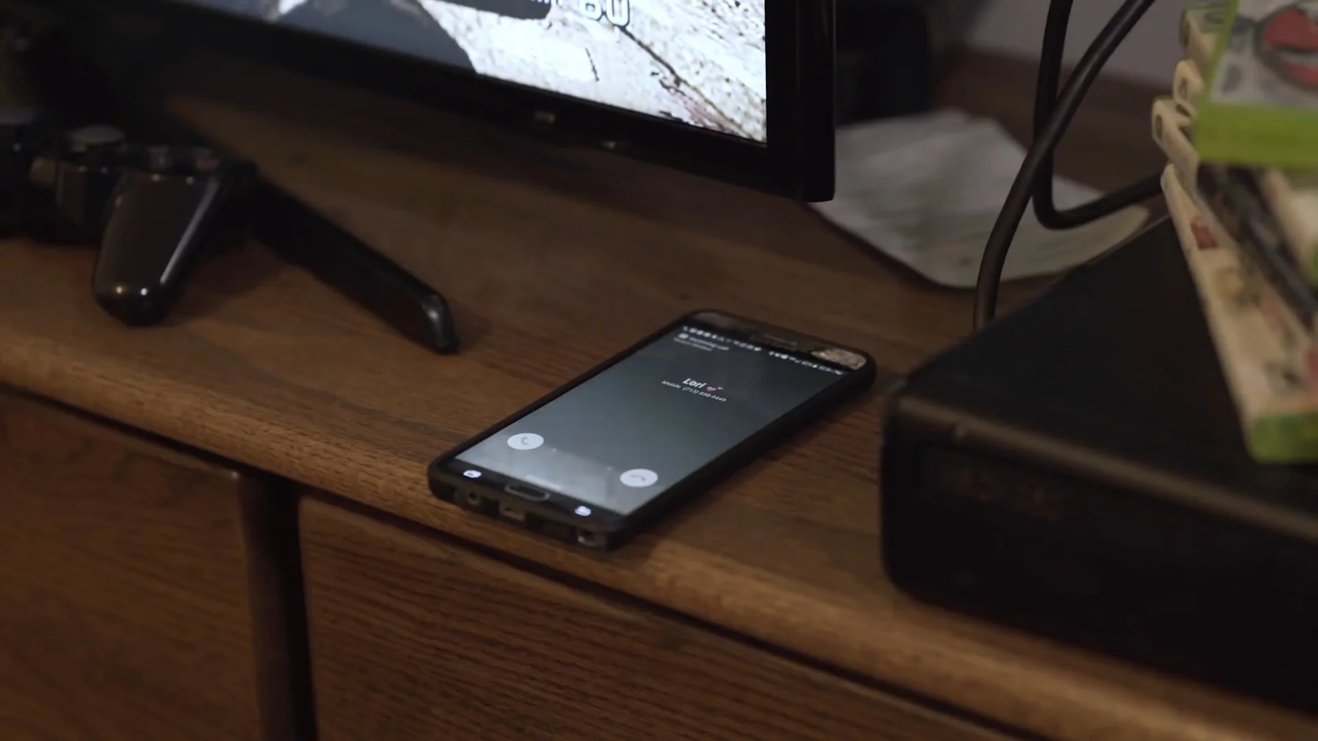 A smartphone displaying an incoming call from 'Loni' resting on a wooden surface, with a TV screen in the background.
