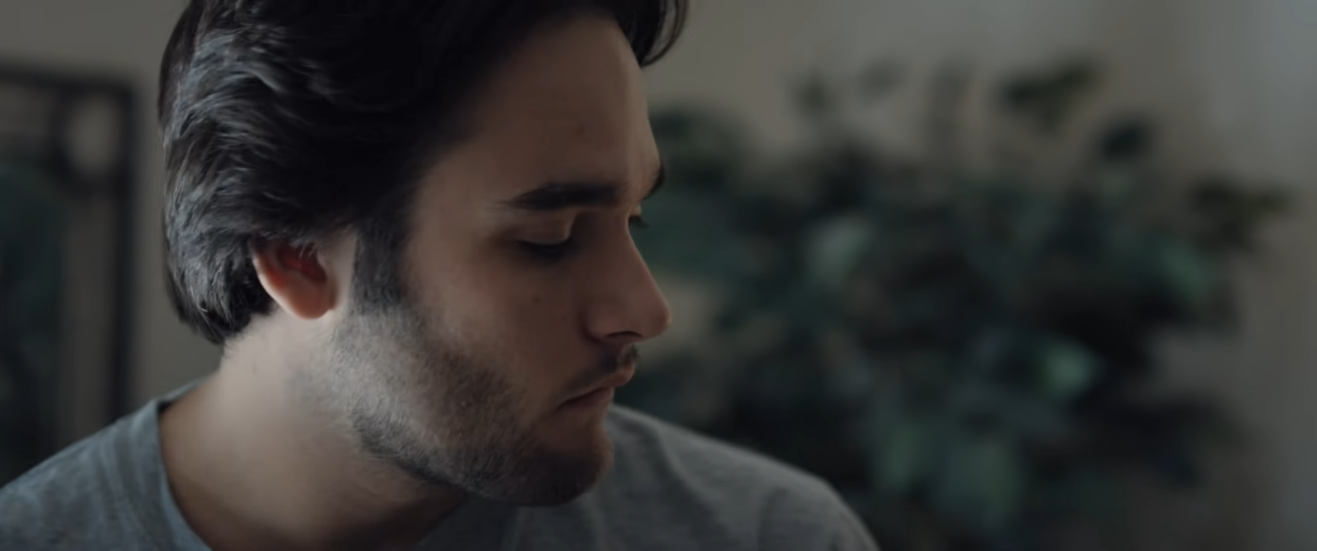 Close-up profile of a young man with dark hair, looking down, with a blurred plant in the background.