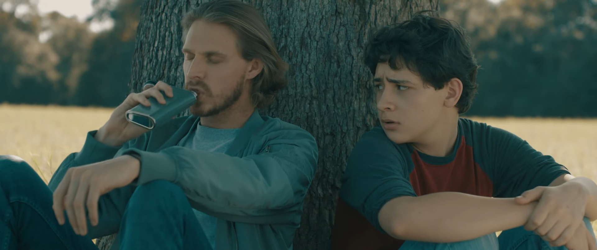 Two young men sitting against a tree, one with long hair drinking from a bottle, the other looking confused or concerned.