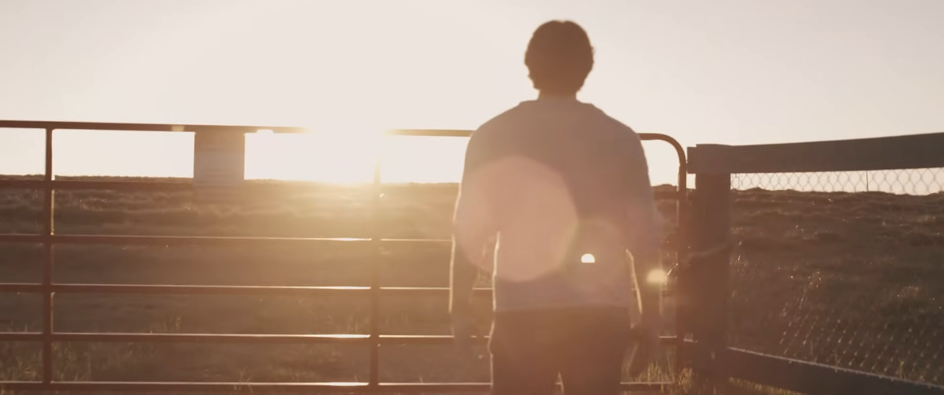 Person standing near a fence, facing away, with the setting sun in the background.