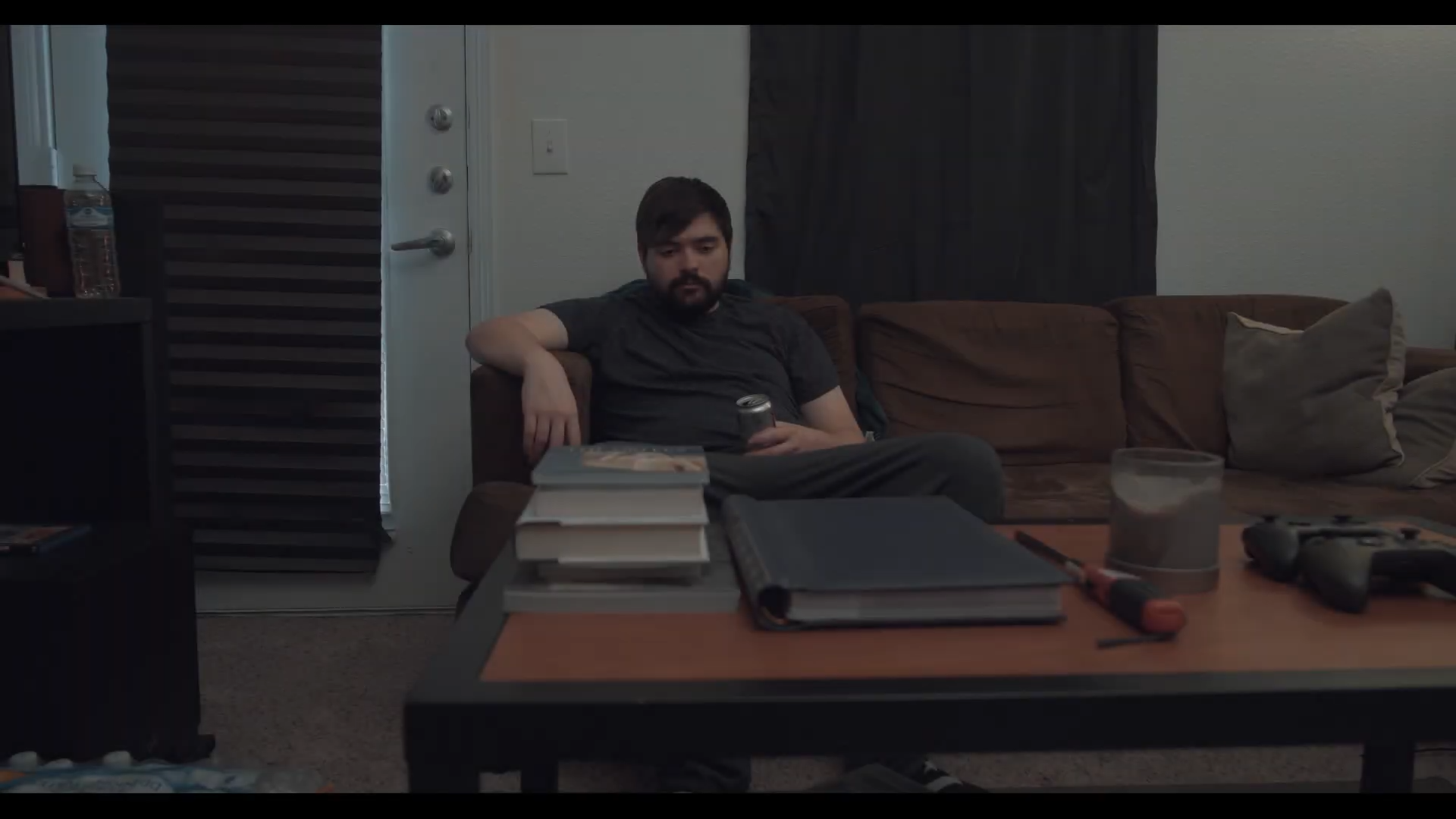 A man sitting on a brown couch in a living room, holding a soda can. There are books, a notebook, and a controller on the table in front of him.