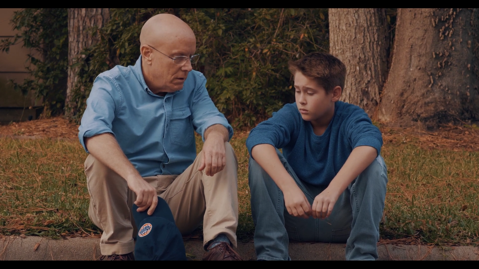 An older man and a young boy sitting on a curb with trees and grass in the background, appearing to be in a serious conversation.
