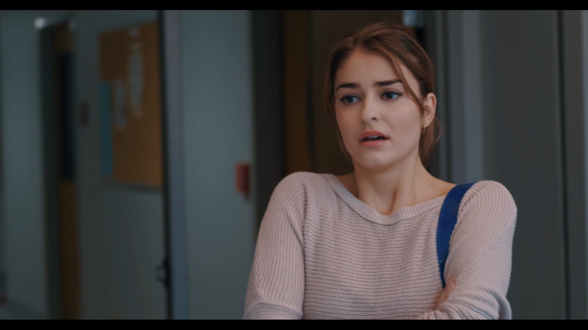 A young woman with shoulder-length brown hair and light skin, wearing a beige sweater and carrying a blue backpack, stands in front of a row of lockers in a school hallway, with a worried or anxious expression on her face.