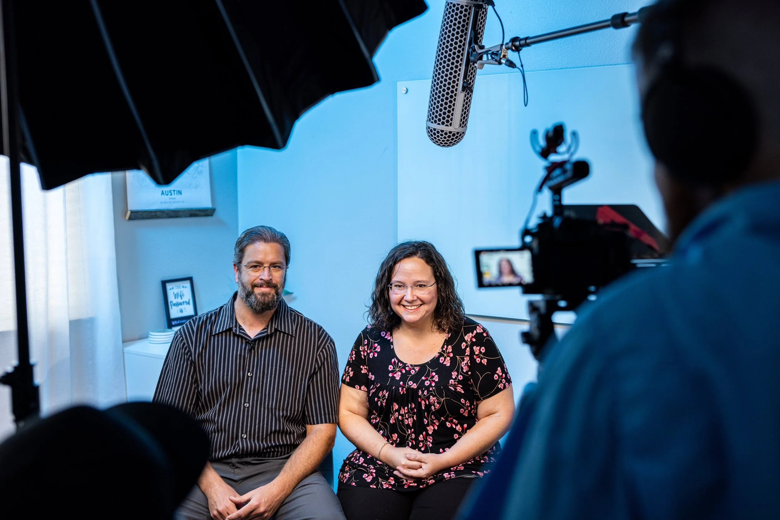 A man and woman sitting side by side during an interview or recording, with filming equipment and a camera person in a blue-walled room.