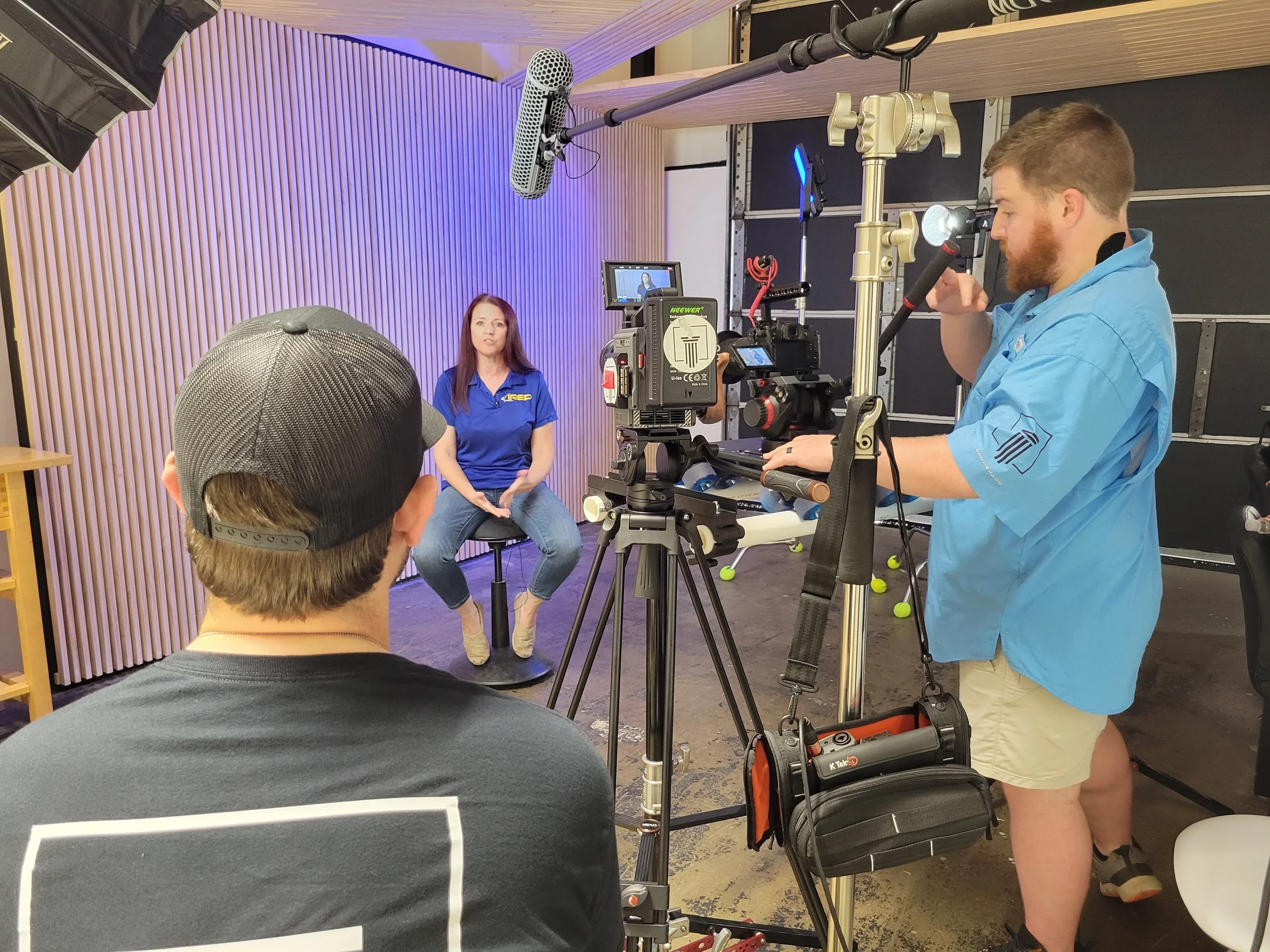 A woman wearing a blue shirt is seated in front of cameras and lighting equipment in a studio setting, with a crew filming her.