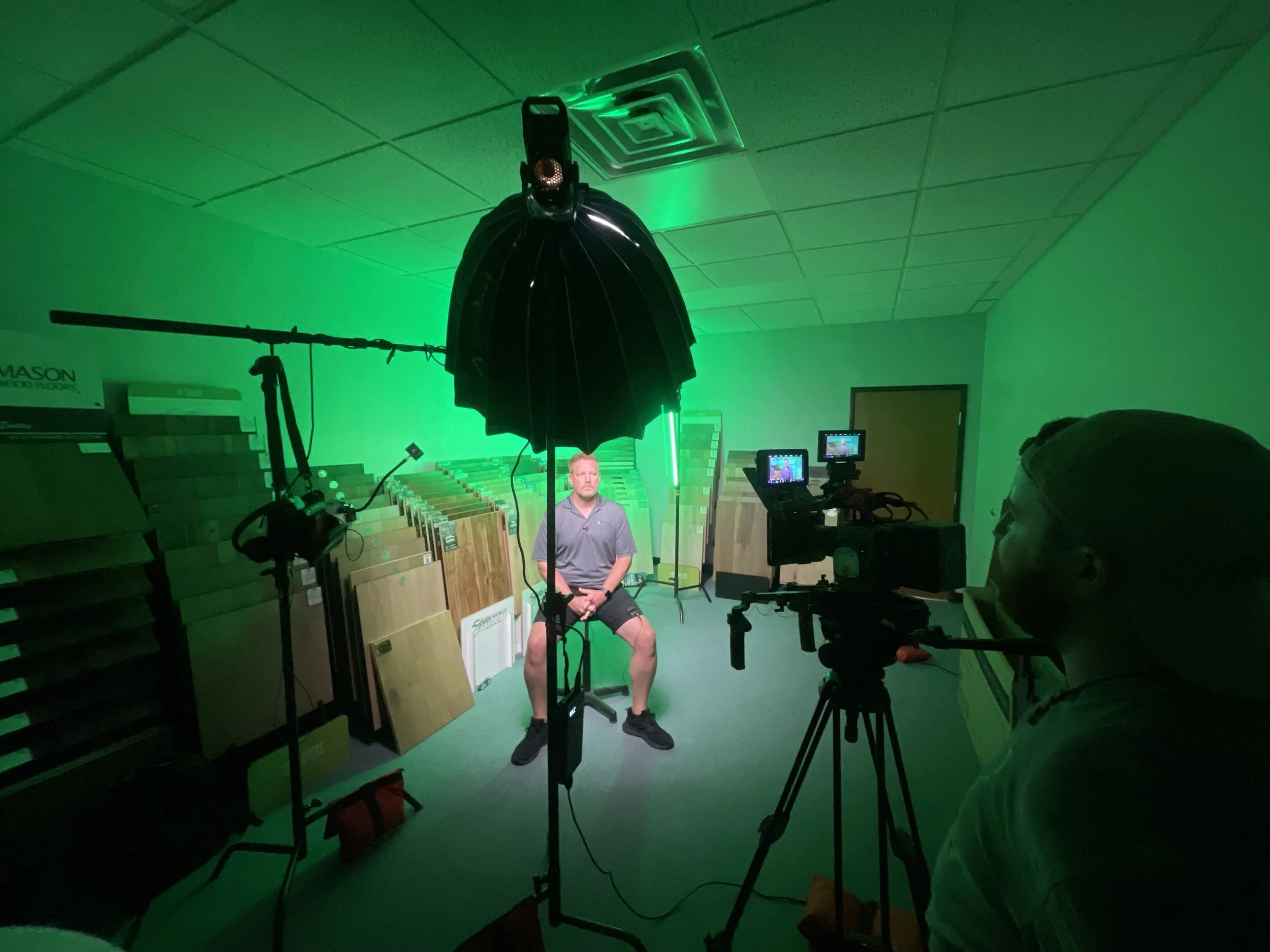 A man is sitting on a stool in front of camera equipment during a video shoot with green screen background, with boxes of flooring samples on either side.